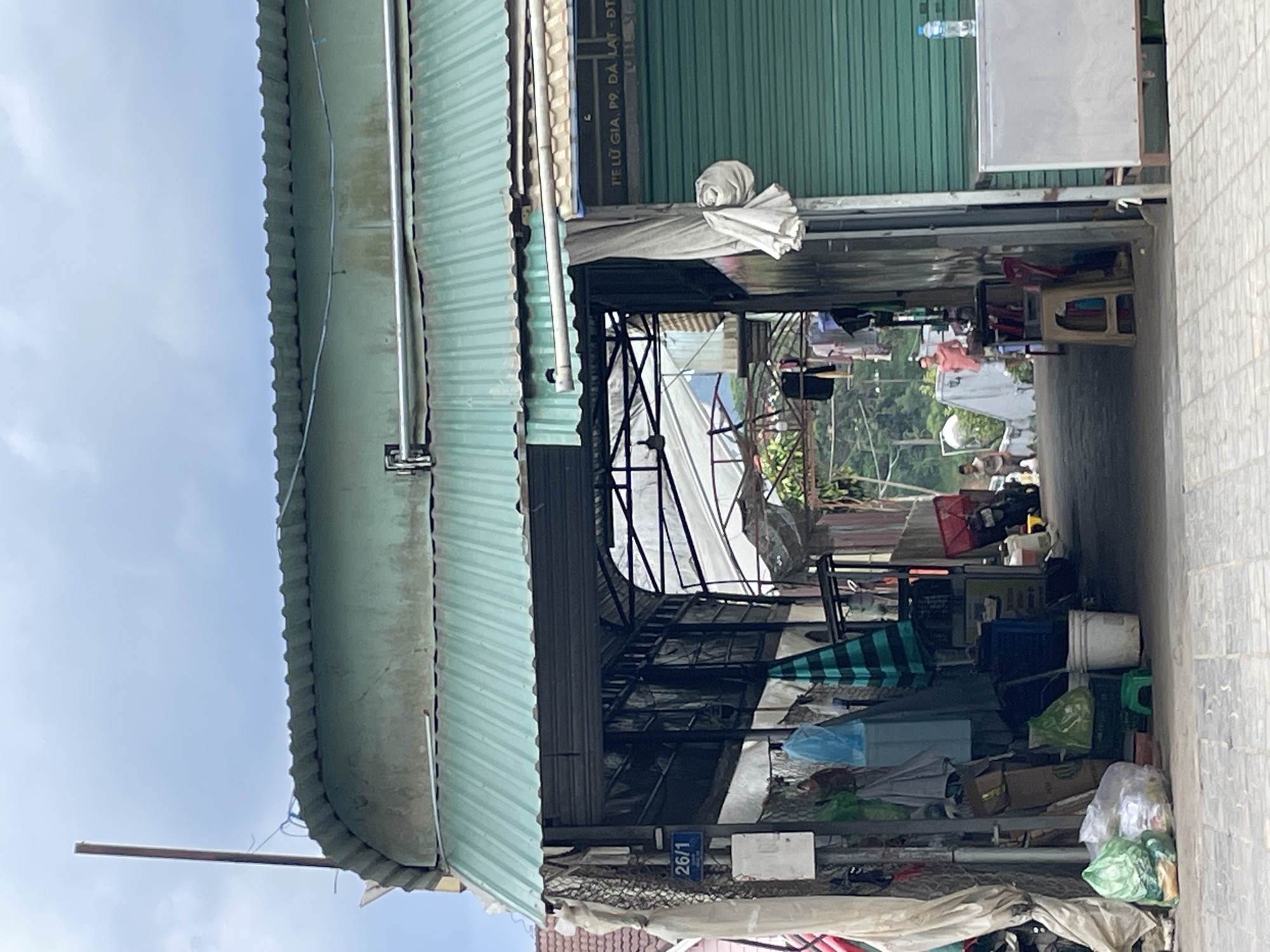The image captures a street scene in Lâm Viên - Đà Lạt, Vietnam, featuring a covered walkway or alleyway with an overhead green corrugated metal roof. On the left side, a blue sign with "26/1" is visible, along with various items stored beneath the roof, including plastic bags of trash, boxes, and hanging clothing. The walkway extends into the background, revealing more of the covered structures and a glimpse of a lush green hillside beyond. Two individuals are seen walking in the distance within the alley. On the right side of the frame, a closed green shuttered storefront is present. The ground is paved with light-colored square tiles. The sky is overcast.