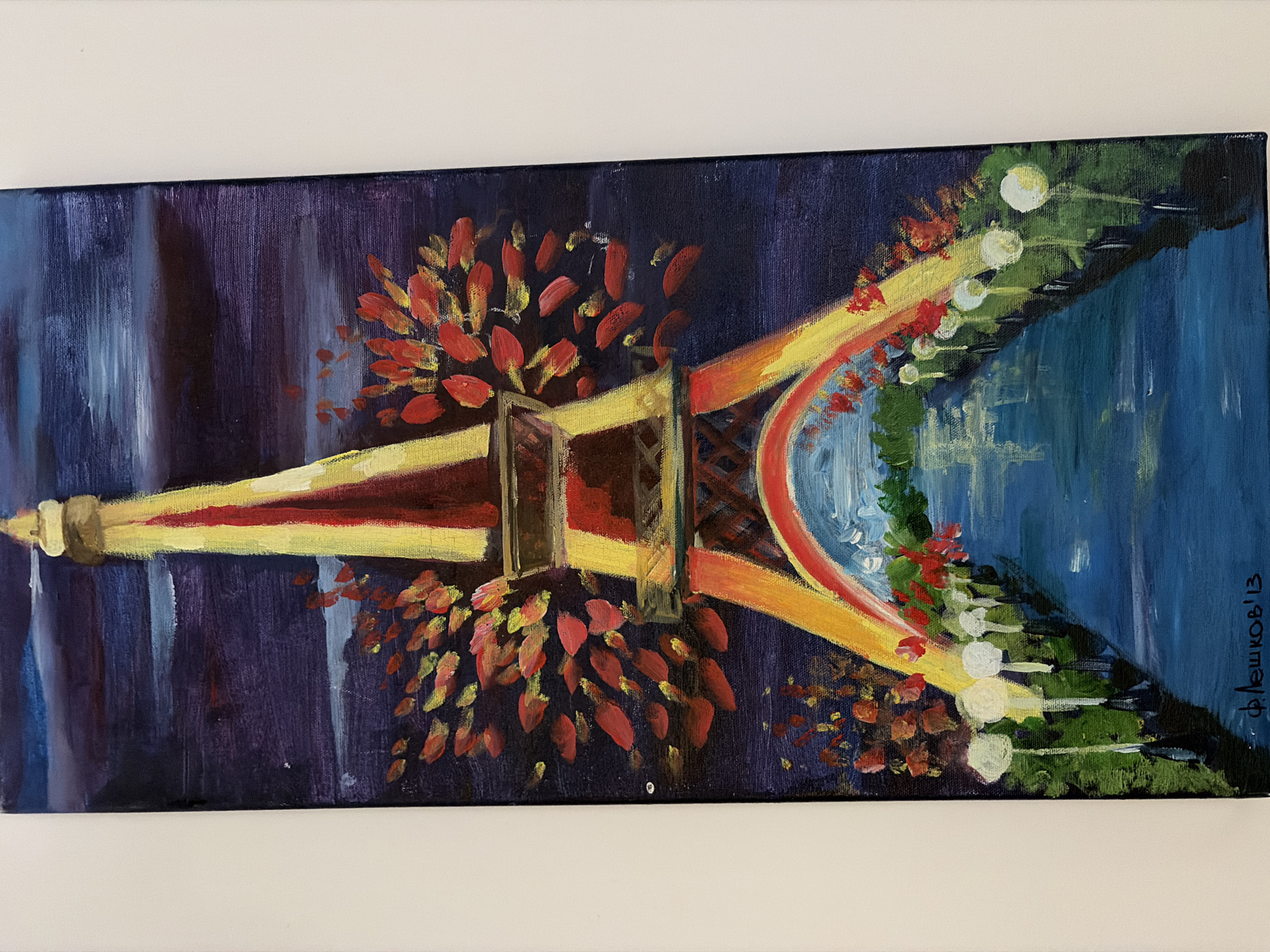 The image is a painting of the Eiffel Tower at night, illuminated by warm yellow and red lights. The tower is depicted against a dark purple and blue sky. Red and orange foliage appears to be blooming from the sides of the tower. In the foreground, a pathway lined with white lights leads towards the tower, with a body of water on either side. The painting is signed in the bottom left corner with Cyrillic script and the year '13. The painting is displayed on a white wall. There is no indication that this scene is located in the city of Germasogeia Municipality, Cyprus.