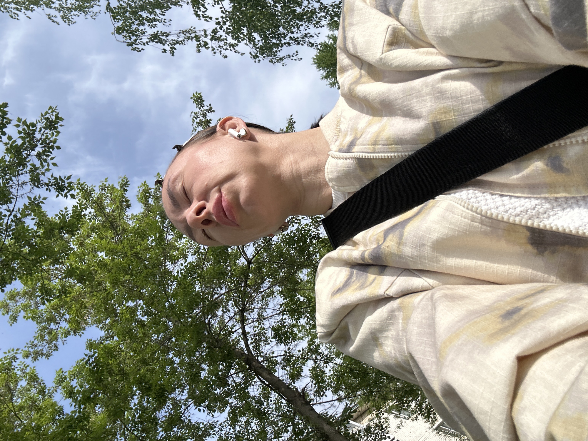 A low-angle shot captures a person with fair skin, an earbud in their left ear, and a closed-eye expression. They are wearing a light-colored jacket with a white, textured interior and a black strap across their torso. Above and around them are green leafy trees against a backdrop of blue sky with scattered clouds. The location appears to be outdoors, although specific details identifying it as Votkinsk, Russia, are not discernible from the image alone. The focus is on the individual and the natural environment surrounding them.