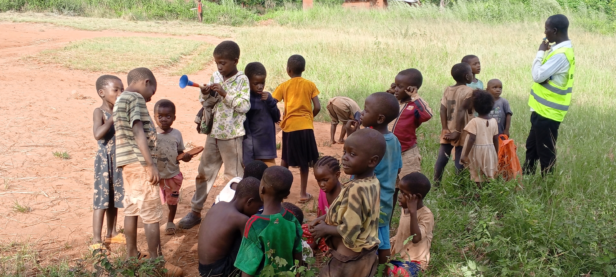 In an outdoor setting near Ngalula, Democratic Republic of the Congo, a group of children are gathered on an area of mixed red dirt and patchy grass, with taller grass growing on the right side of the frame. There are approximately twenty children of varying ages, mostly facing different directions. One child is holding a blue and white horn-like instrument. Another is holding a sandal. A man is standing off to the right in the taller grass wearing a white shirt, black pants, and a bright yellow reflective vest. He appears to be talking or observing.
