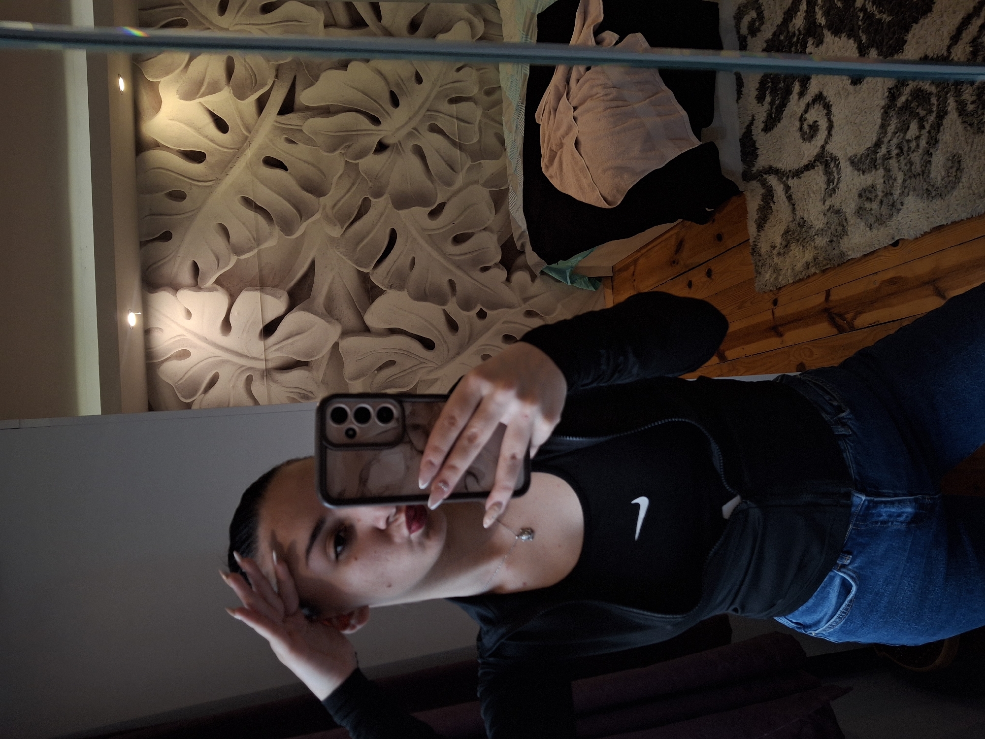 A young woman is taking a selfie in a mirror. She is wearing a black athletic top with a white Nike swoosh and blue jeans. She has long nails and a silver necklace. The background includes a wall with a large tropical leaf pattern, a rug, and wooden flooring. It is not possible to confirm the location is Buhovo, Bulgaria from the image.