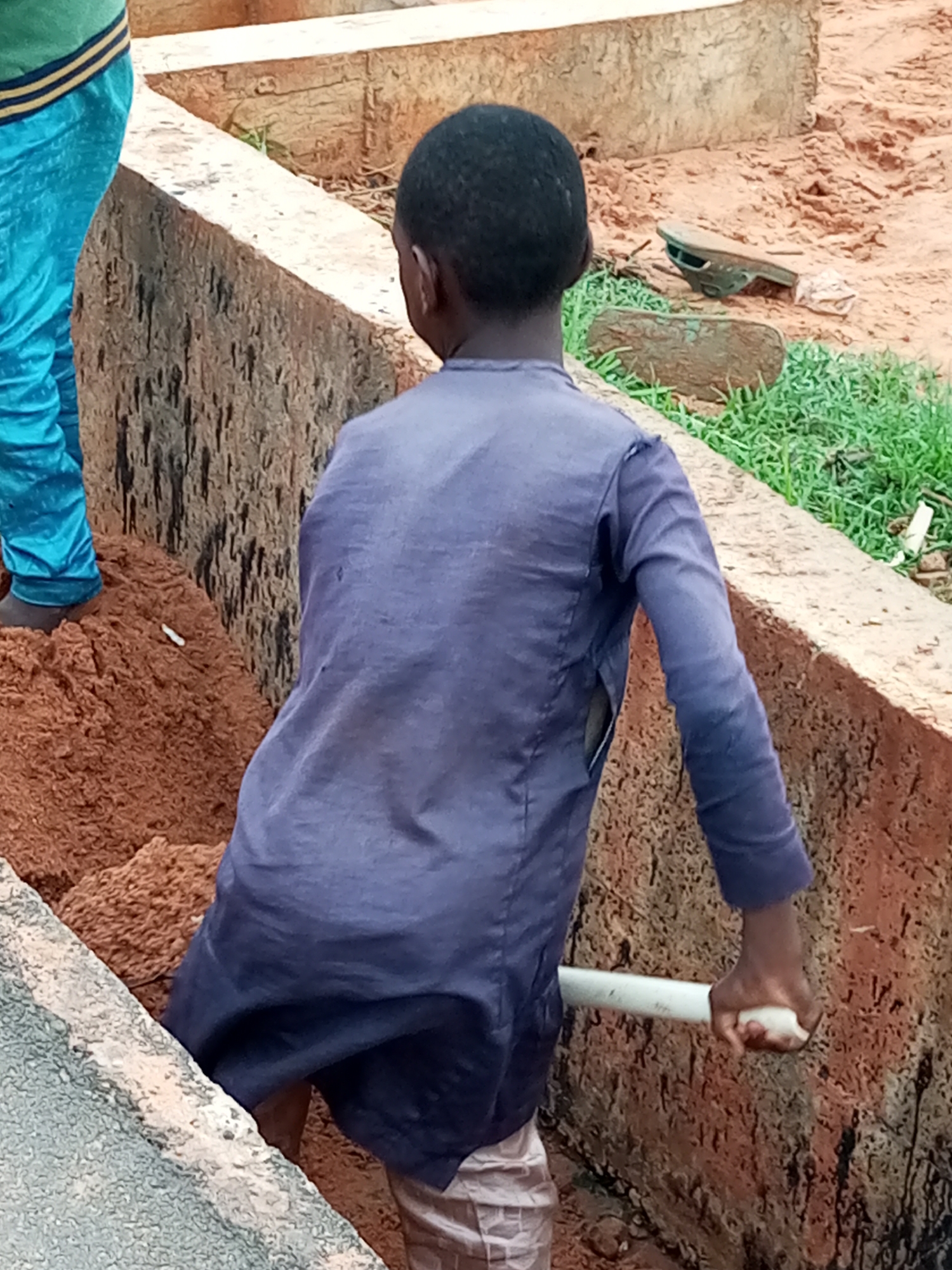 The image shows a young boy in a blue long-sleeved shirt and plaid pants digging in a construction area. He is holding a white pipe in his right hand, as if using it as a tool to move dirt. To his left, another person wearing bright blue pants and a green and yellow striped shirt is partially visible. The ground is covered in reddish-brown dirt and some green grass is growing along the edge of a concrete structure. The overall environment appears to be an outdoor construction or excavation site.