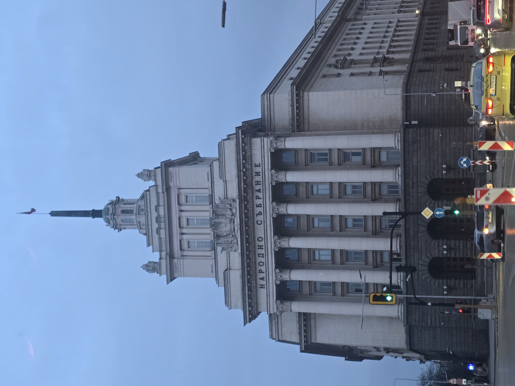 The image shows the National Assembly of Bulgaria building in Sofia. The building is a large, symmetrical structure with a classical design, featuring columns and a central spire. The words "НАРОДНО СЪБРАНИЕ" (National Assembly) are inscribed on the facade. A yellow taxi with the license plate "CB 7179 HX" and other cars are on the street, indicating traffic. Traffic lights are visible, and the sky is overcast. The environment appears to be urban, with standard road signs and street furniture.