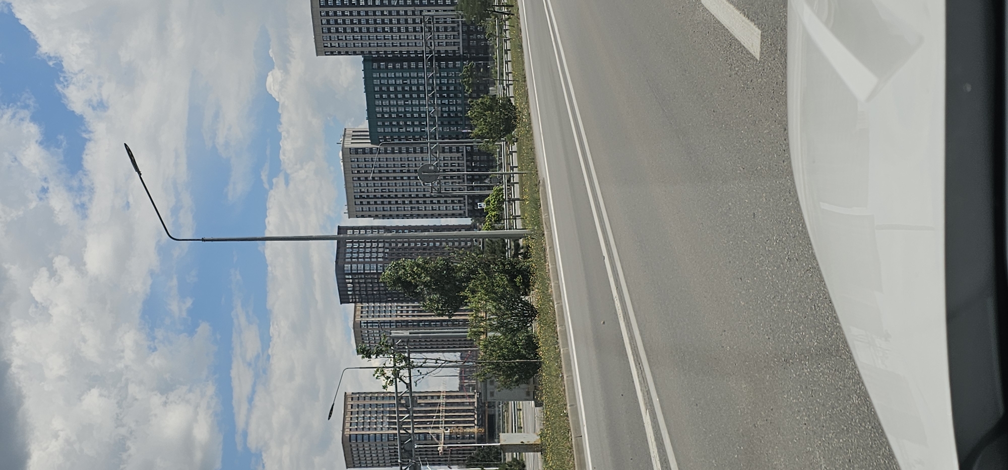 The image shows a street scene in the city of Овощной, Russia. In the background, there are several multi-story residential buildings. The buildings are modern in design, with numerous windows and balconies. In the foreground, there is a paved road with white lane markings. A white car is visible at the bottom of the frame, suggesting the photo was taken from inside a vehicle. There are streetlights and some vegetation along the side of the road. The sky is partly cloudy.