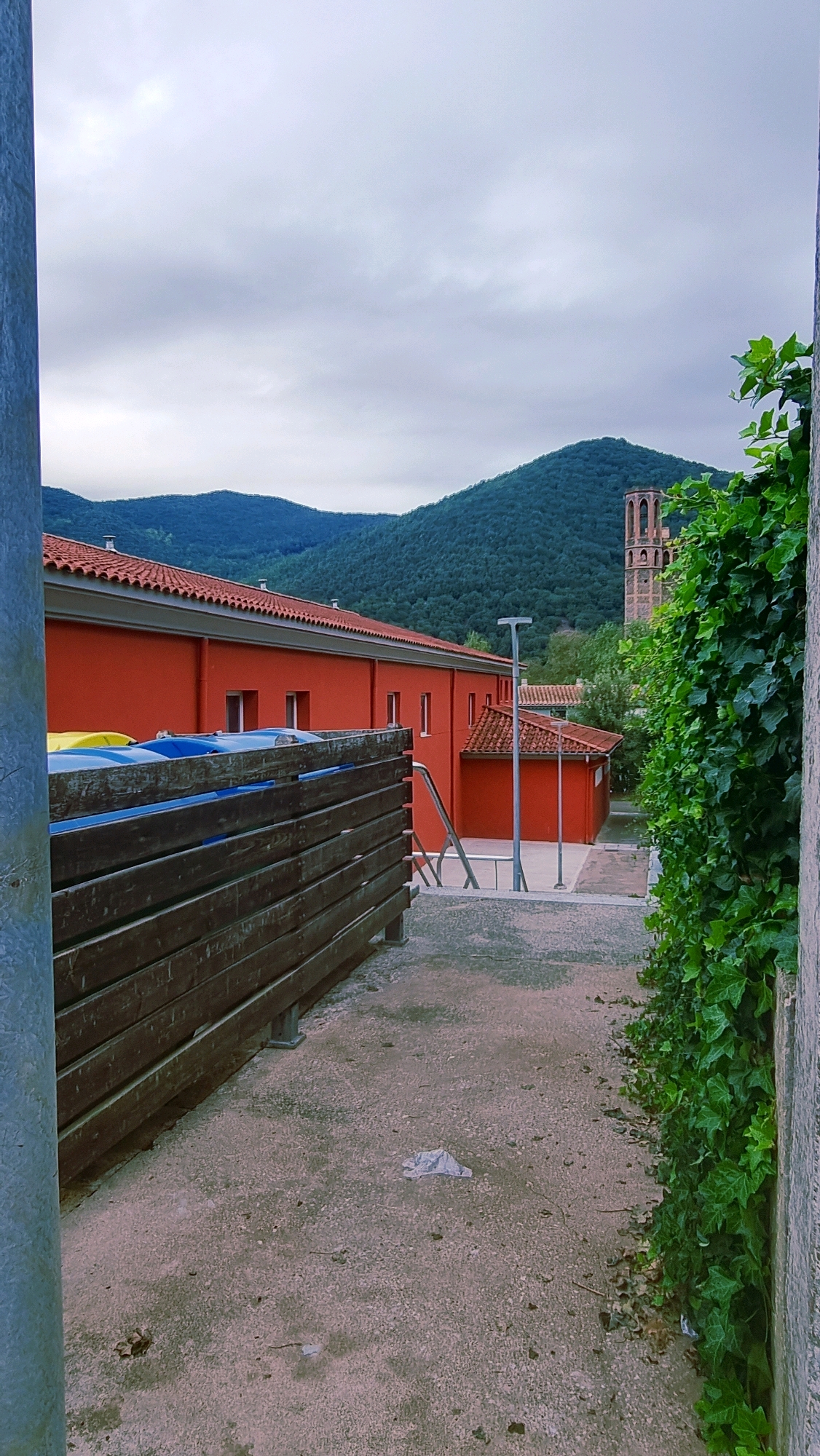 The image depicts an outdoor scene in Sant Joan Les Fonts, Spain, under an overcast sky. In the foreground, a rough wooden fence runs along the left side of a paved pathway. The pathway is a pale gray concrete, showing signs of wear and some scattered debris. To the right of the pathway, a dense wall of green ivy climbs up a textured, light-colored wall.
Dominating the mid-ground is a vibrant red building with a terracotta-tiled roof. The building has a series of uniformly spaced windows. Adjacent to this, a smaller red structure with a similar roof is visible, and a metal lamp post stands between them. To the far right of the mid-ground, a tall, ancient-looking stone tower with arched openings rises above the trees, suggesting a historical or religious site.
The background is characterized by rolling, verdant hills covered in dense forest, their shapes softened by the atmospheric conditions. The overall impression is one of a tranquil, somewhat rural setting within the town. No people are visible in the frame.