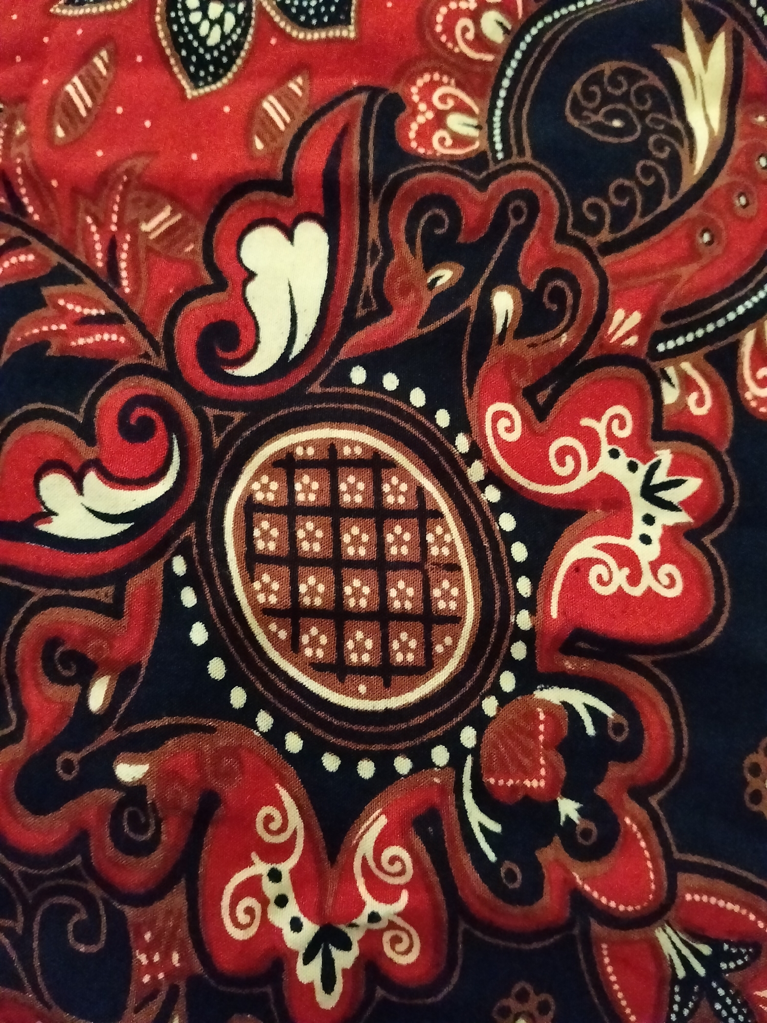 A close-up shot captures a detail of a red, black, and cream batik fabric. The pattern features intricate floral and swirling motifs. In the center, an oval shape contains a grid of small squares, each with a white dot. Surrounding this central element are red petal-like shapes with cream accents and delicate white swirls. Dark blue areas form the background and outline some of the patterns. The texture of the fabric is visible, suggesting a woven material. There are no people or specific landmarks visible in this image, therefore its location in Gempolan, Indonesia cannot be determined from the image alone.