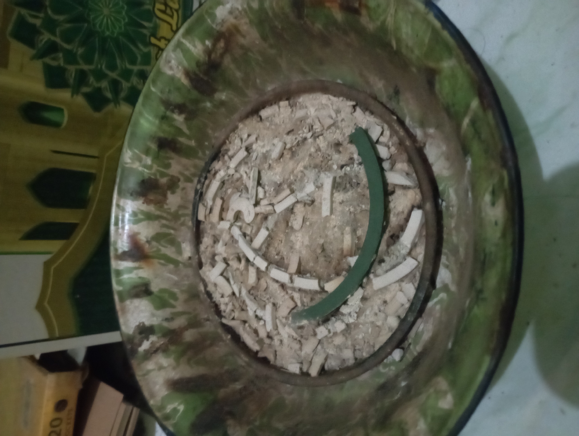 An overhead close-up shot focuses on a dirty, round, metal ashtray filled with cigarette butts and ash. A green mosquito coil is partially burned in the center of the ashtray, with some segments still intact and others broken. The ashtray itself has a marbleized pattern of green and brown. In the background, out of focus, is a box with the number "20" visible, and a hint of a green decorative pattern, possibly from a wall or banner, with arch-like shapes. The scene is lit from above, casting a shadow at the bottom of the frame. The location is identified as Sembung Anyar, Indonesia.