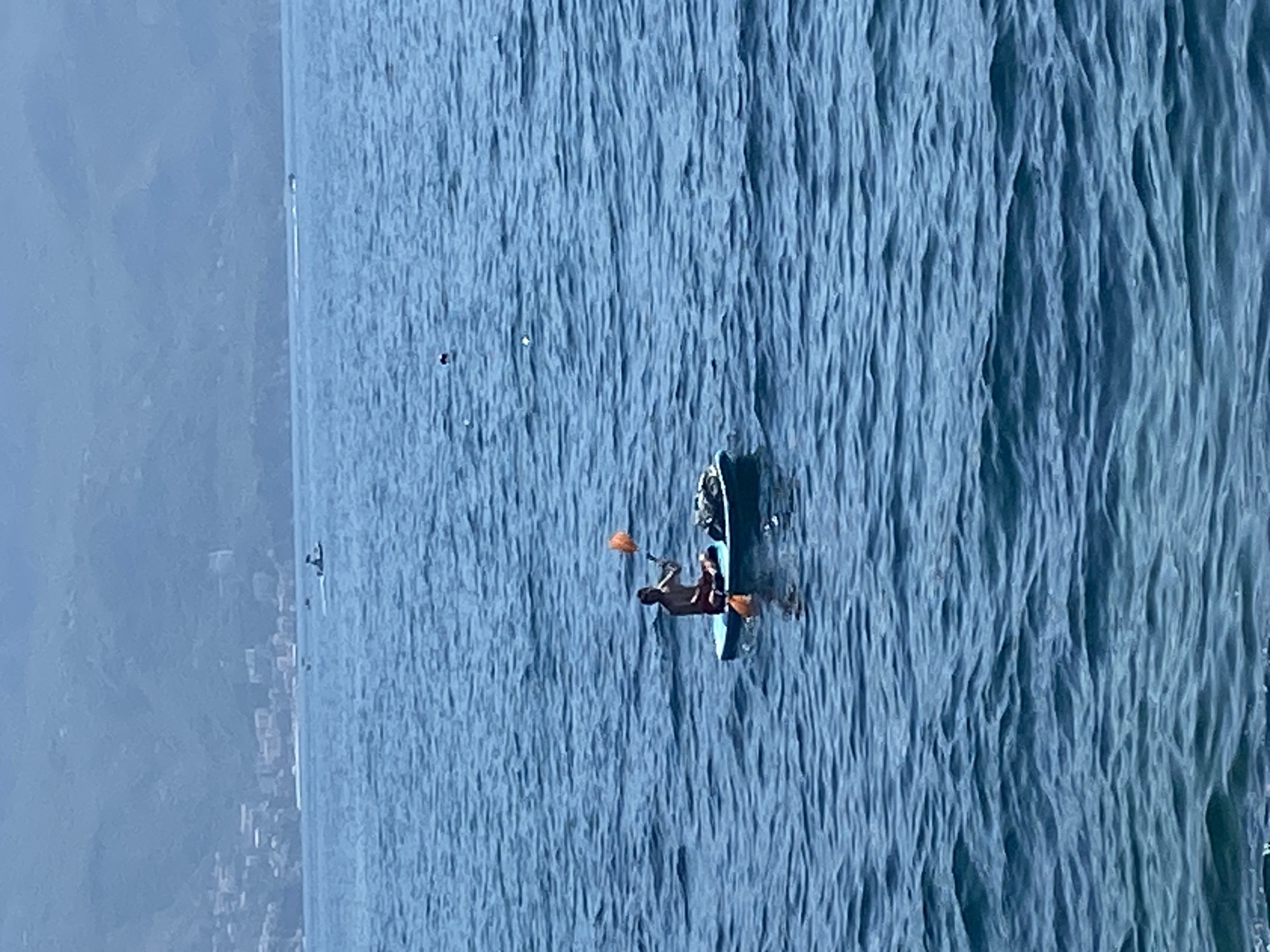 A person is paddleboarding on a blue sea with a mountain range in the background. The person is sitting on the paddleboard and holding an orange object. There is a bag on the paddleboard. In the distance, there are buildings on the coast and a few boats on the water. The sea is choppy with small waves. The sky is overcast. The scene appears to be in Budva, Montenegro.