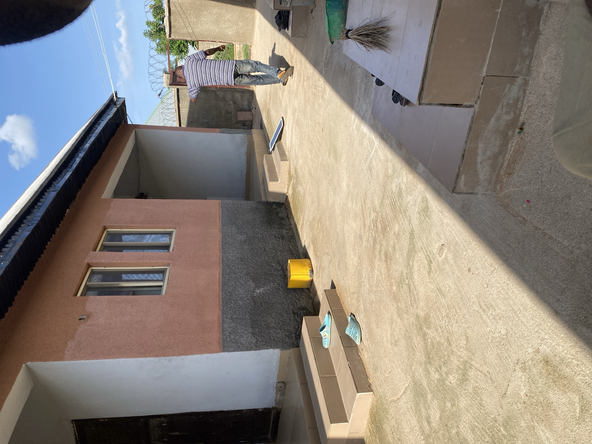 In Jauro Yinu, Nigeria, a man in a blue and white striped shirt and blue jeans walks away from the camera in a courtyard. He is walking towards a chain-link fence topped with barbed wire, which encloses the property. To the left of the man, there is a single-story building with a coral-colored facade and a dark gray lower section. The building has two windows with light-colored frames. In front of the building, there are two steps with light brown tile risers. On the upper step, a pair of blue slip-on shoes are placed, and on the lower step, another pair of blue slip-on shoes is visible. A bright yellow cylindrical object is located to the right of the steps. A long, rectangular solar panel lies flat on the ground in the middle of the courtyard. To the right of the courtyard, a dark gray concrete wall stands, and further to the right, a light brown tiled platform is visible with a bundle of dry grass and a green plastic basin on it. The ground in the courtyard is concrete, with visible wear and some discoloration. The sky is blue with scattered white clouds.