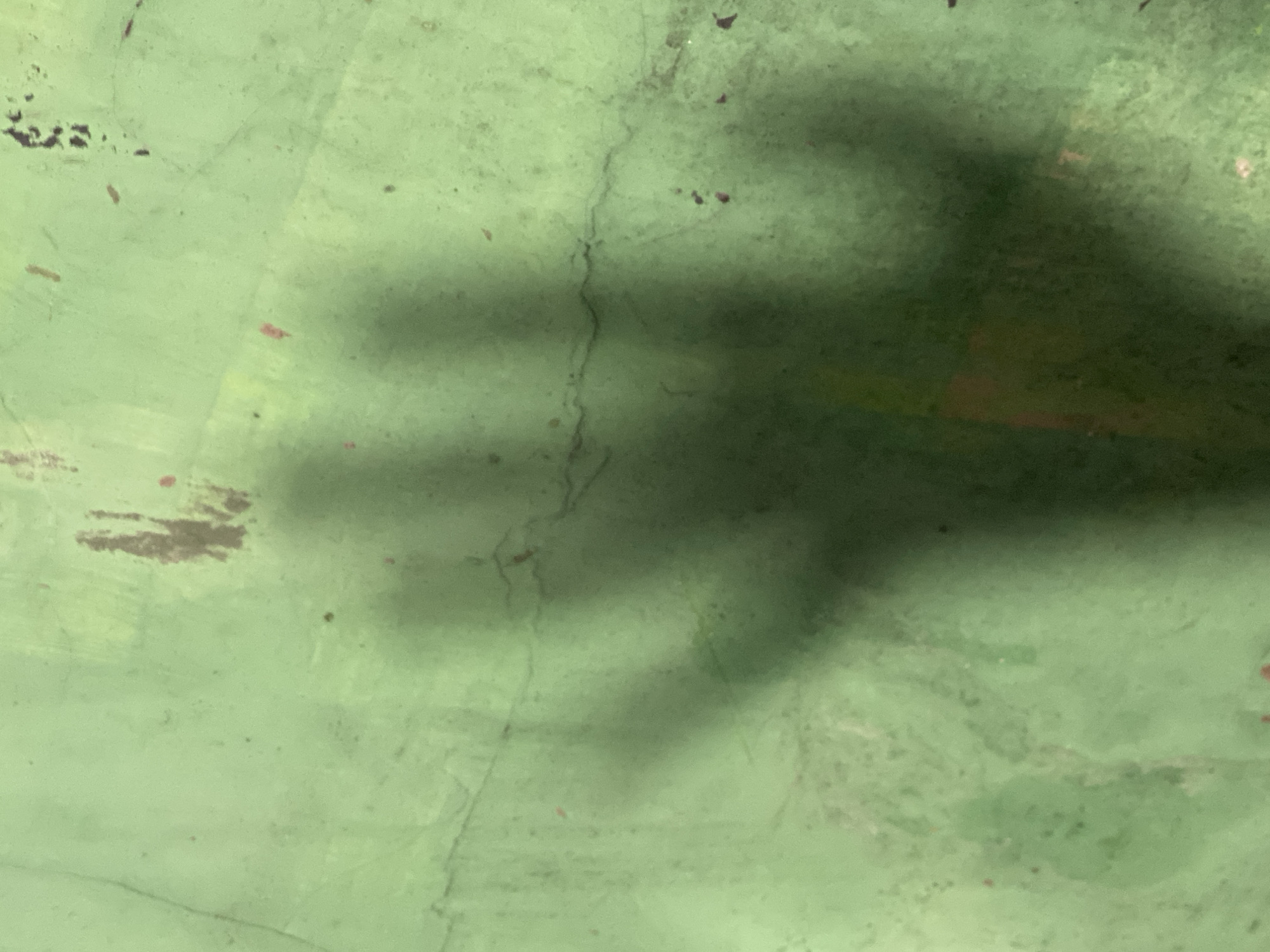 The image is a close-up, full-frame shot of a shadow cast on a green, textured surface, possibly a wall or ground. The shadow, which is dark and slightly blurred, resembles a hand with spread fingers. The green surface has visible cracks and some reddish-brown stains, suggesting it may be an outdoor or weathered area. The lighting indicates the shadow is cast by an overhead light source. The image was captured in Kasihan, Indonesia.