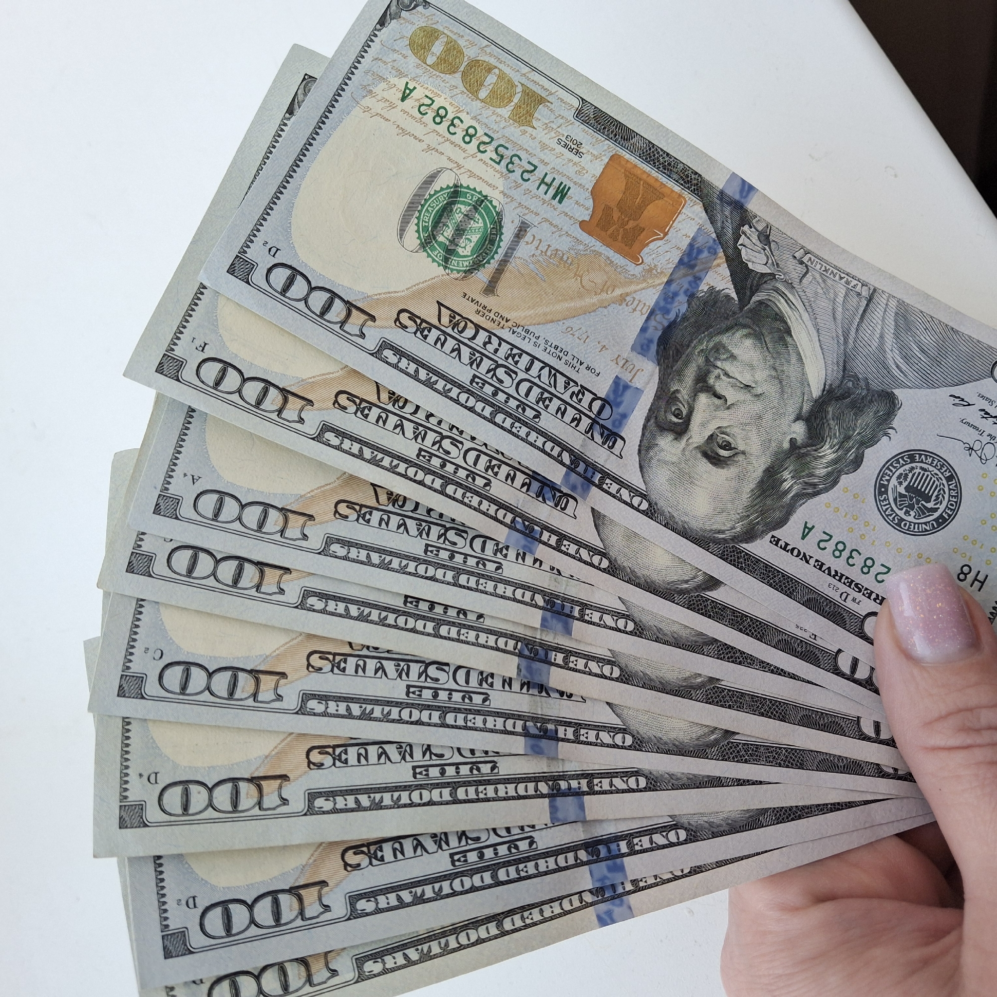 A close-up shot shows a person holding a fan of United States one hundred dollar bills. The bills are arranged in a cascading fashion, with the top bill partially obscuring the others. The hand holding the bills is visible, showcasing a manicured nail with a sparkly design. The background is a plain white surface.
