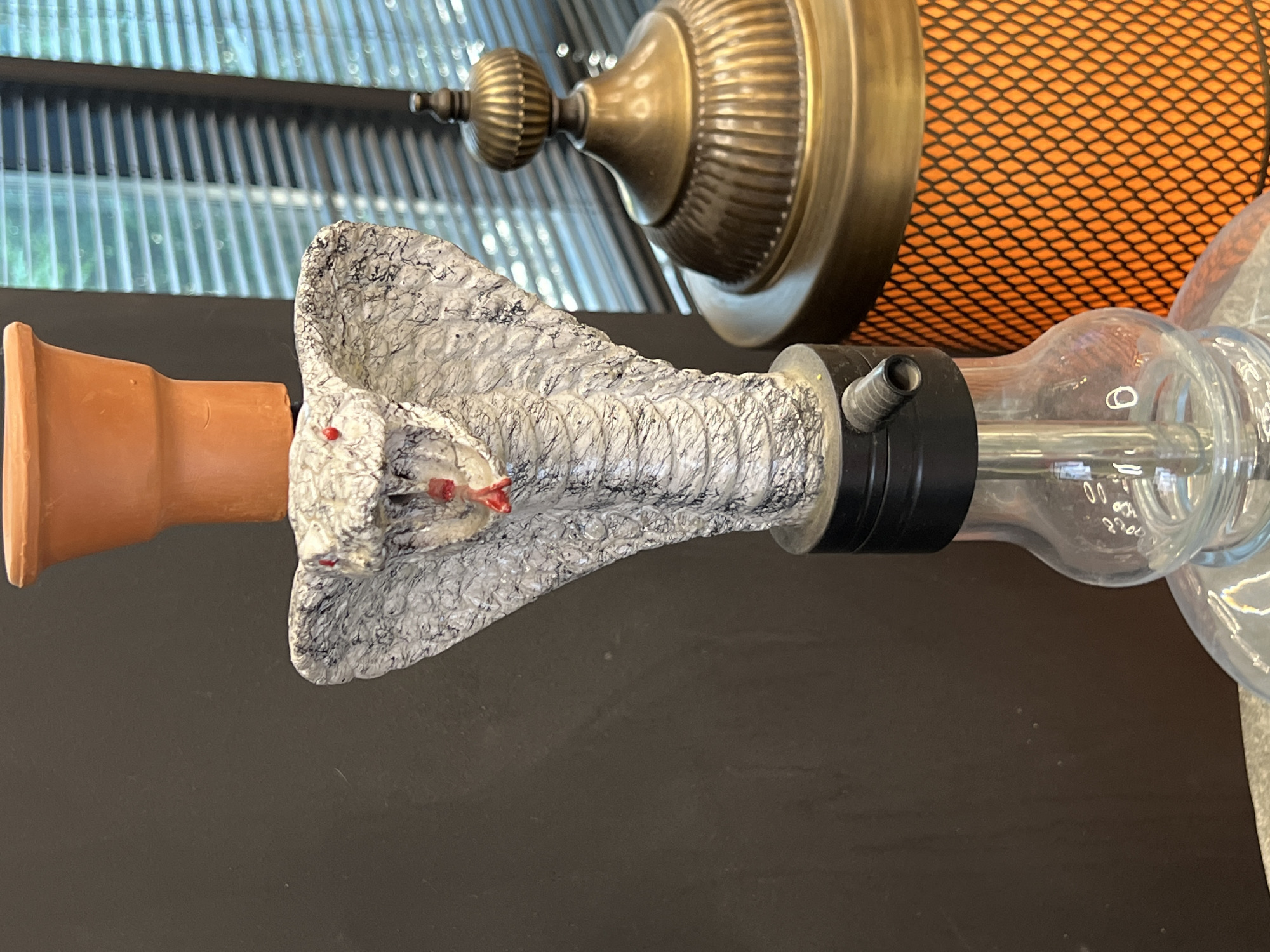 The image shows a close-up of a hookah's pipe and bowl. The pipe is designed to resemble a cobra's head, with a textured, gray and black patterned body and small red accents on its face, possibly representing eyes or fangs. The cobra's head is attached to a clear glass tube, which is part of the hookah's body. A black rubber seal connects the cobra head to the glass. To the left of the cobra head is a reddish-brown, cylindrical object, likely the hookah's bowl, made of what appears to be terracotta. In the background, there is a ribbed, translucent surface, possibly a window or skylight, and a glimpse of a decorative brass lamp with an orange, mesh-like shade. The scene is indoors, with artificial lighting and a dark surface in the foreground. No people are visible, and the focus is on the intricately designed hookah components. The setting appears to be a room or display area where such items are kept or sold. There is no visible text in the image.