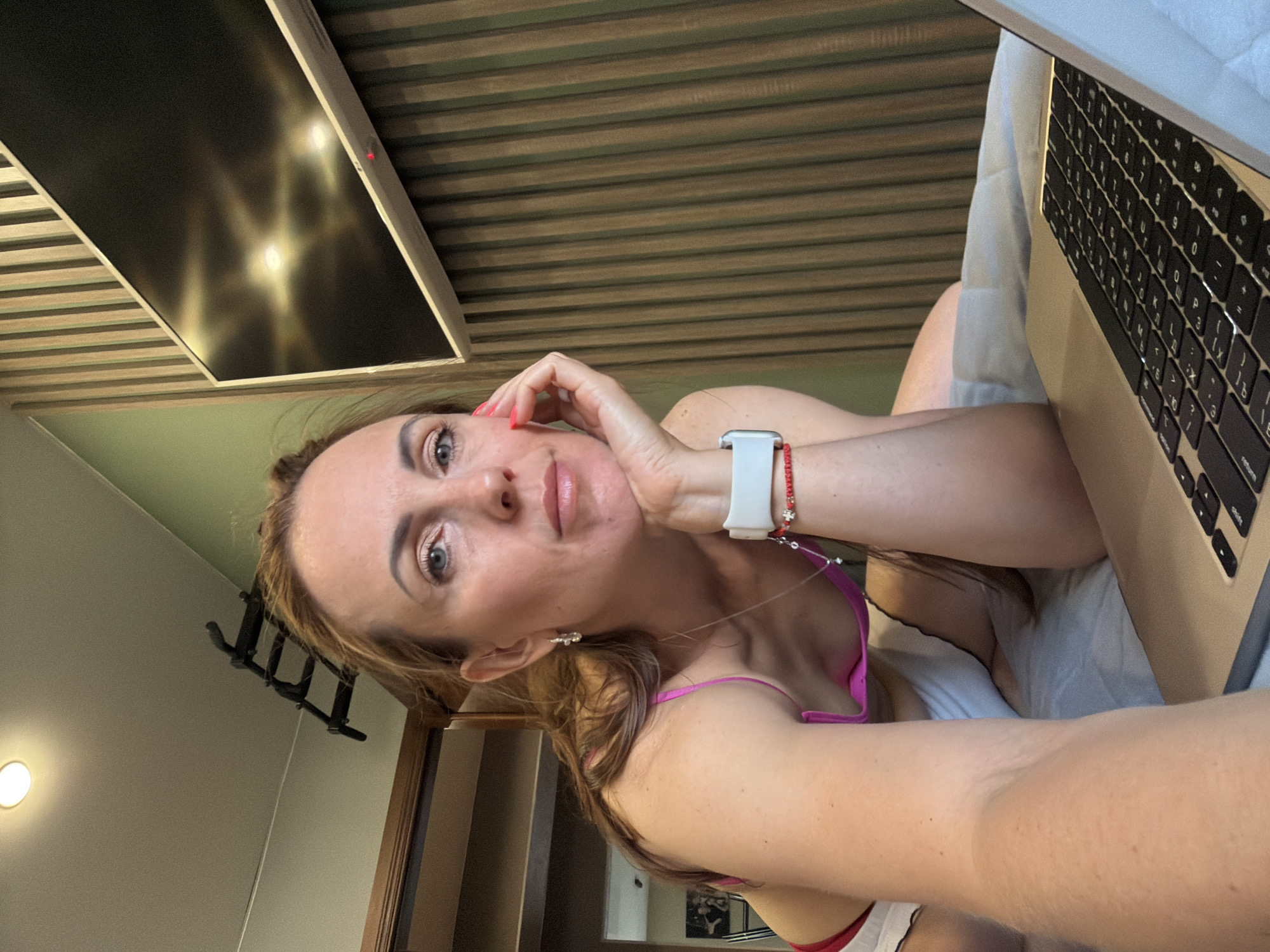 The image shows a woman with light brown hair and blue eyes, wearing a pink top and white shorts. She is resting her head on her hand, looking towards the camera. A laptop is in front of her, and a television is mounted on the wall behind her. The room appears to be a bedroom, with a bed and a ceiling light visible. The walls are decorated with vertical wooden slats. The overall impression is that of a casual, indoor setting, possibly in a home or hotel room. There is no indication of the location being in Birobidzhan, Russia, or any specific city.