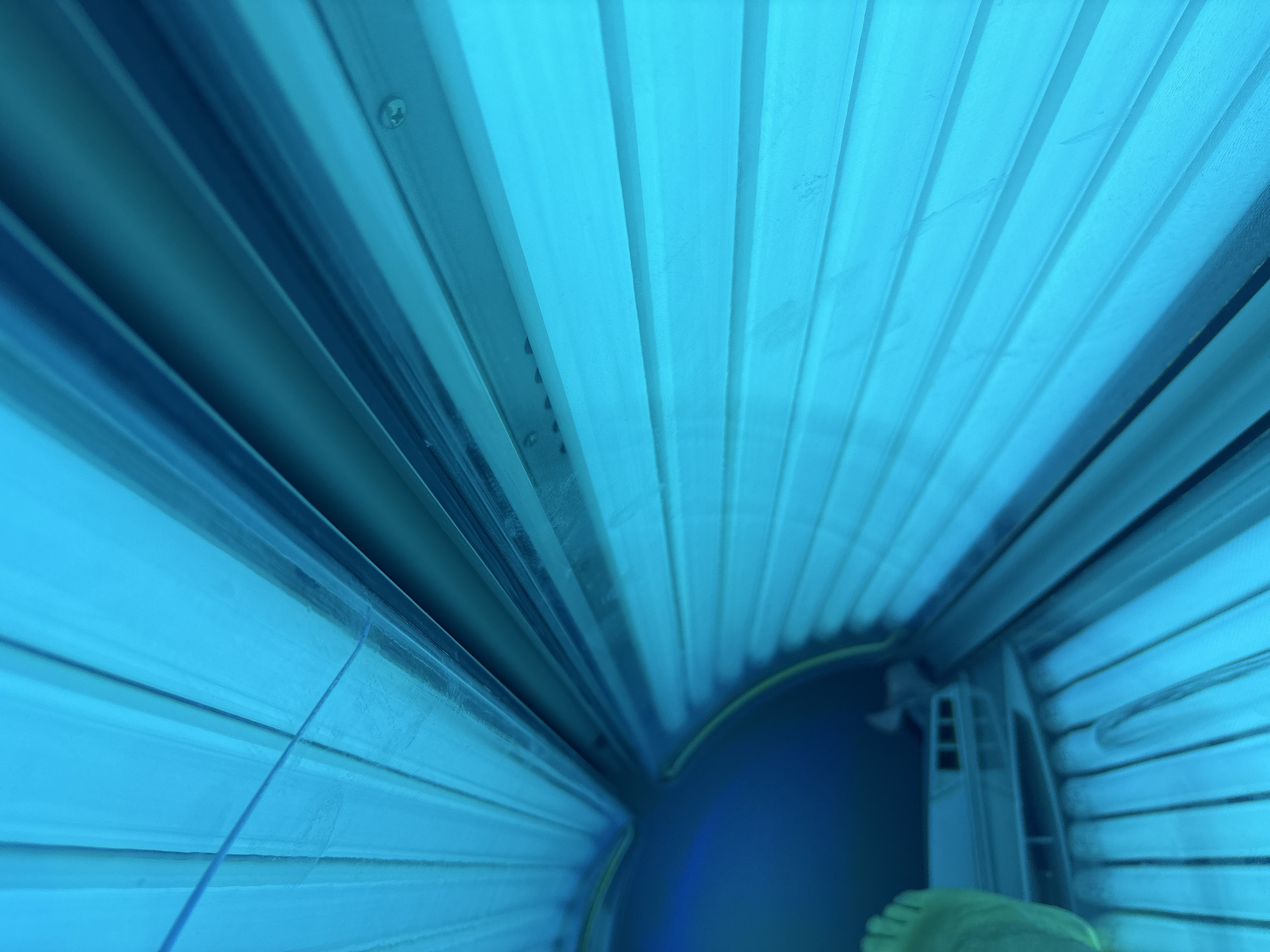 The image shows the interior of a tanning bed. The tanning bed is illuminated with a blue light. The interior consists of horizontal blue panes. There are several screws visible. There is a foot in the lower right corner. The image does not contain enough information to determine the location in Varna, Bulgaria.