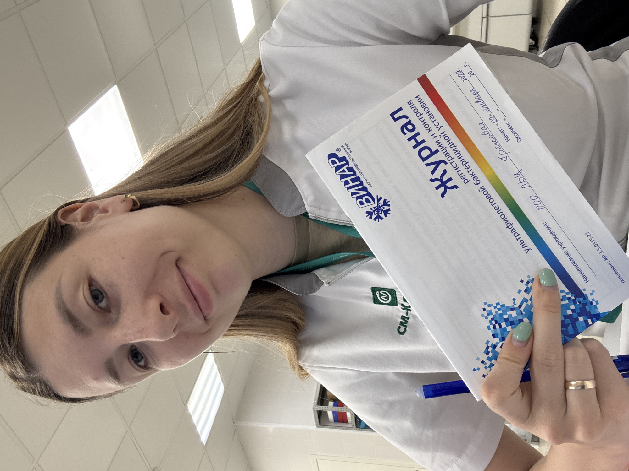 A young woman, likely in a professional setting such as a medical facility or laboratory, is captured in a close-up, upward-angled shot. She is wearing a white lab coat or medical uniform, under which a green lanyard is visible. A "CM-K" logo is embroidered on her uniform. She has long, light brown hair, fair skin with some freckles, and light-colored eyes. Her expression is calm with a slight smile, and she is looking directly at the camera.
She is holding a white document, identified as a "Журнал регистрации и контроля ультрафиолетовой бактерицидной установки" (Journal of registration and control of ultraviolet bactericidal installation), and a blue pen. The document features the logo of "ВИНАР" (VINAR), a "НАУЧНО-ПРОИЗВОДСТВЕННАЯ ФИРМА" (Scientific-Production Company). Handwritten details on the document include "ООО ЛВЧ Грекова" (LLC LVC Grekova) and "Начат: «02» января 2026г." (Started: "02" January 2026). Her nails are painted light green, and she wears a simple gold ring on her left ring finger and a small gold hoop earring on her visible ear.
The background shows a brightly lit indoor space with white ceilings featuring grid patterns and fluorescent light panels. Parts of shelves with items are visible behind her. The overall setting, combined with the uniform and the document's content, suggests a clinical or scientific environment, consistent with the provided location context of Moscow, Russia.