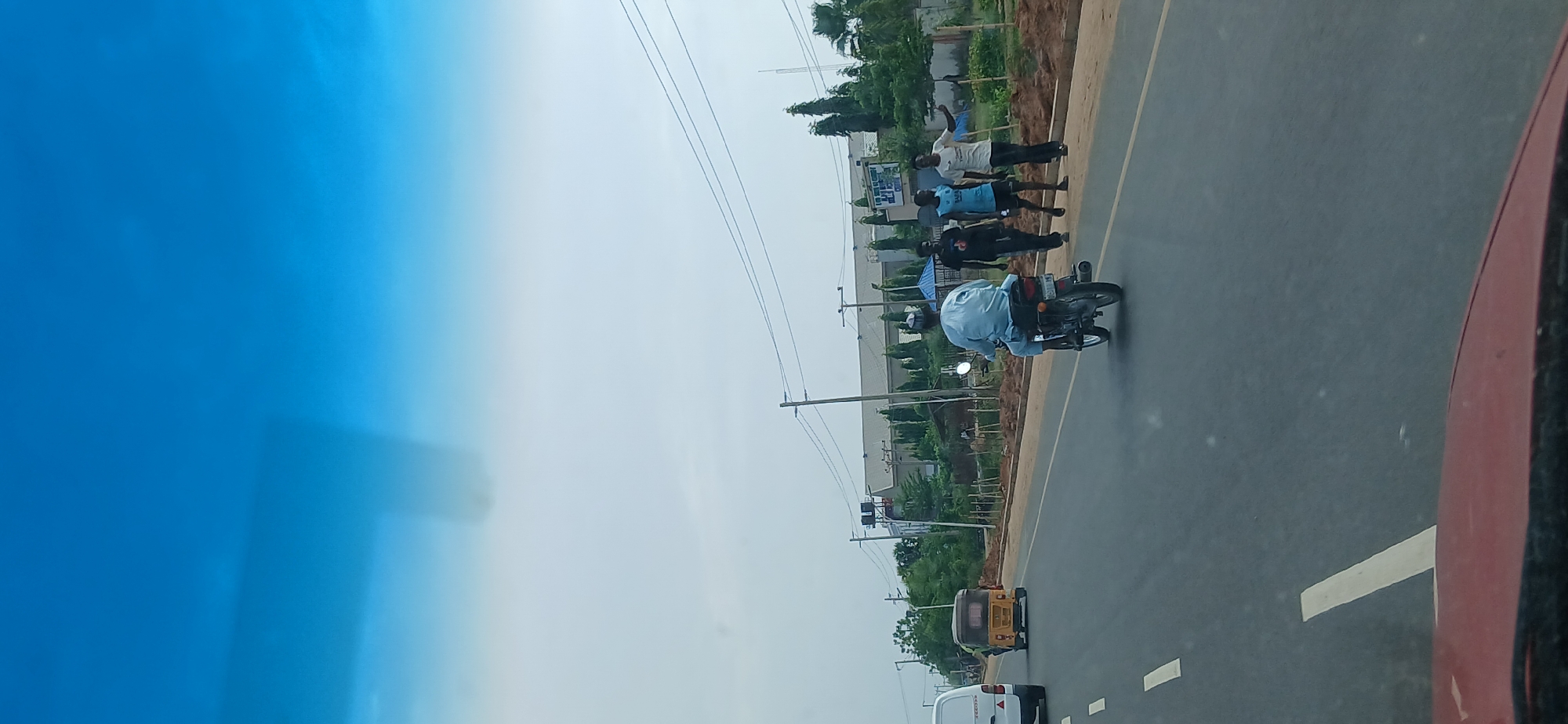 The image shows a view from inside a vehicle, possibly a car, looking out onto a road in Matage, Nigeria. The weather appears overcast. On the right side of the road, a group of men is standing near a grassy shoulder. A motorcyclist is driving on the road. Further down the road are an auto rickshaw and a white van. Electrical poles and wires are visible overhead. The blue hue in the upper left corner indicates the windshield of the vehicle from which the photo was taken.