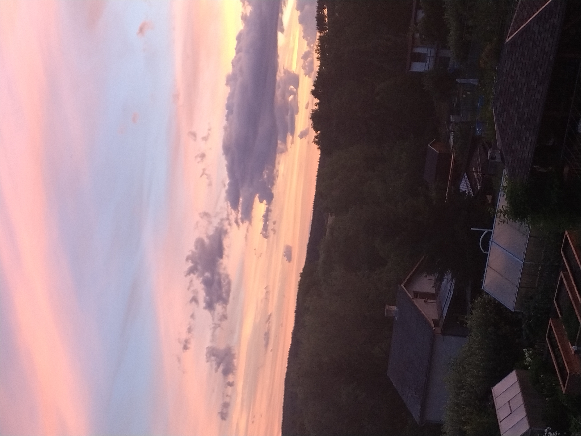 The image is a photograph taken in Dobev, Czechia, capturing a rural landscape at dusk. The sky is filled with a dramatic sunset, displaying soft pinks, purples, and oranges, with scattered dark clouds. Below the sky, a dense line of dark green trees forms a silhouette against the horizon, suggesting a hilly terrain. In the foreground, several structures are visible. On the left, a house with a dark roof and a chimney is nestled among the trees. To its right, two greenhouses with translucent coverings are situated. In front of the greenhouses, several raised garden beds are visible. Further to the right and slightly in the background, another building with a lighter facade and dark trim can be seen. The overall scene is one of tranquil twilight in a semi-rural or suburban setting.