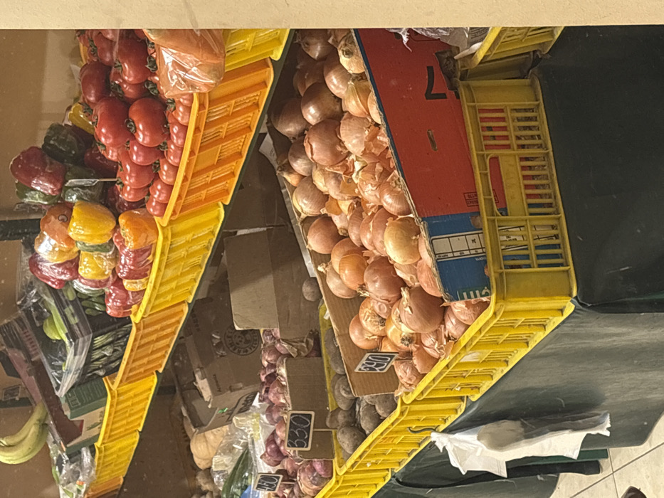 The image displays a static scene within a produce section, located in the city of Unknown, Unknown. Multiple tiers of produce are arranged on shelves, primarily within yellow plastic crates.
On the top tier, from left to right, are bananas in a dark container, green peppers in clear plastic packaging and an open cardboard box, then a large display of red, yellow, and orange bell peppers, many individually wrapped in clear plastic, within yellow crates. To the far right, red tomatoes are piled in yellow crates, with some orange-colored produce, possibly carrots, in a clear plastic bag.
The middle tier features various vegetables, including items in cardboard boxes; one visible box is labeled "Armenia". Red onions are present in a yellow crate, accompanied by a cardboard price tag displaying "330". Adjacent to these are dark-skinned potatoes or similar tubers in another yellow crate. Further right, brown and yellow onions are displayed in a yellow crate, with a partial cardboard price tag showing "290".
The lower section of the display, draped with a dark green fabric, also contains brown and yellow onions, some contained within a red and blue cardboard box inserted into a yellow crate. A white plastic bag hangs from the front of the display on the left. The floor is a light-colored tiled surface. No individuals are present, and no actions or interactions are depicted.