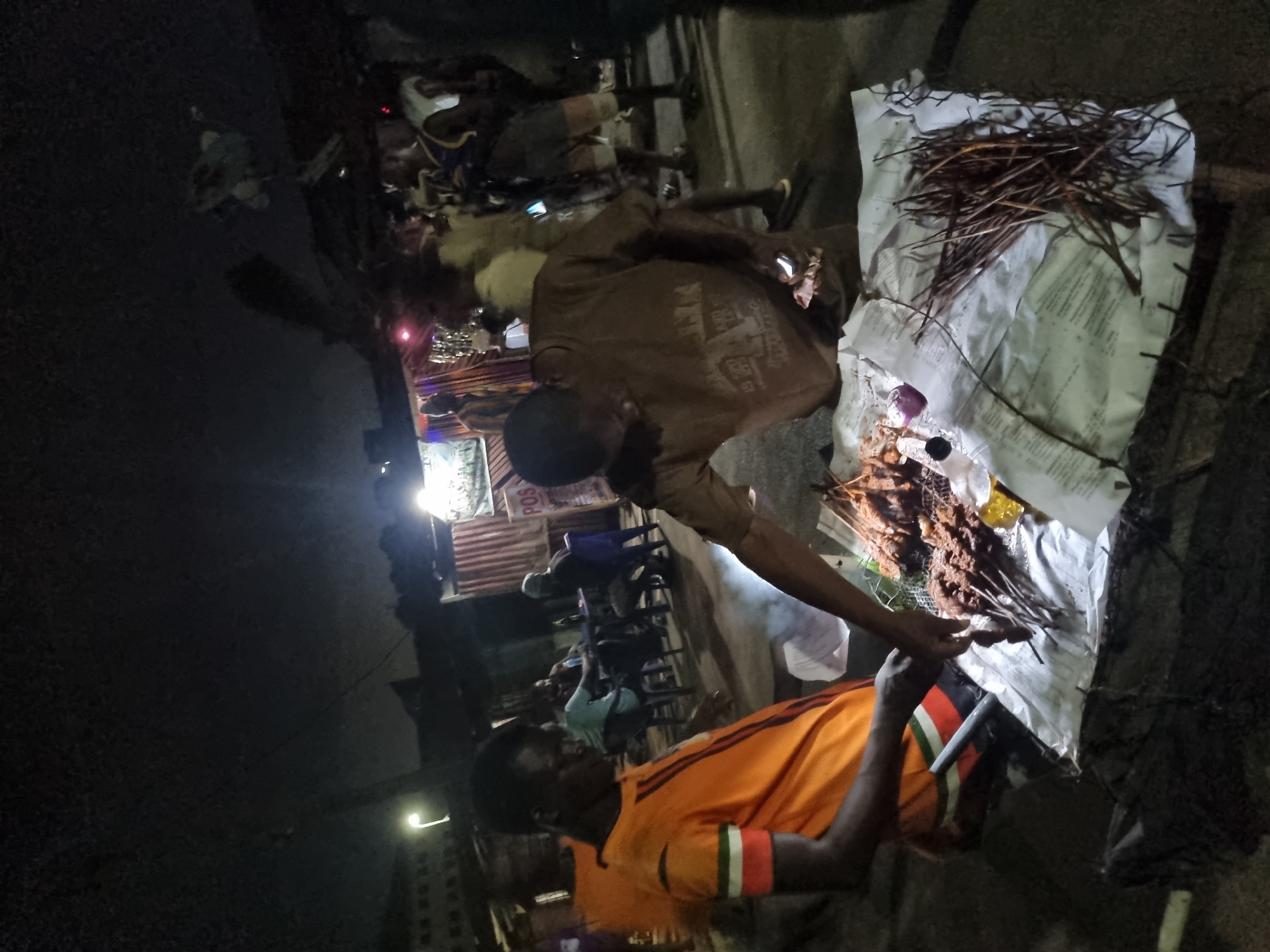The image depicts a late evening street food scene in Onitsha, Nigeria. Two men are visible attending to a charcoal grill laden with suya skewers. One man wears an orange shirt with dark green and white striped sleeves and the other a brown t-shirt. The suya skewers are placed on a makeshift stand covered in white paper or cloth, accompanied by what appear to be sticks or skewers for serving. Behind the men, seated individuals are eating. A partially lit building with signage stands in the background. The location is recognizable as Onitsha due to the ubiquitous street food vending style and general ambiance.