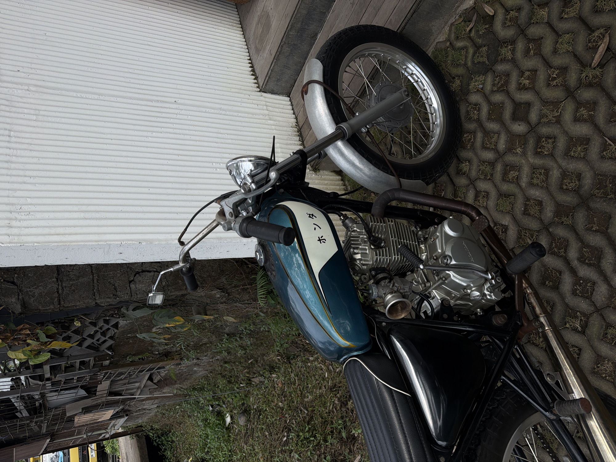 The image shows a custom motorcycle parked outdoors on a patterned concrete surface. The motorcycle has a teal and white gas tank with gold accents and Japanese writing that reads "ホンダ" (Honda). It features a black ribbed seat, a chrome headlight, and silver fenders. The engine is exposed and appears to be a single-cylinder model. In the background, a textured white wall and wooden steps are visible. To the left of the motorcycle, a rough stone wall and overgrown vegetation can be seen. The scene is set in an outdoor environment, possibly in a residential or commercial area. The specific location in Mengwi, Indonesia, is implied but not explicitly shown in the image itself. There are no people or explicit actions depicted.