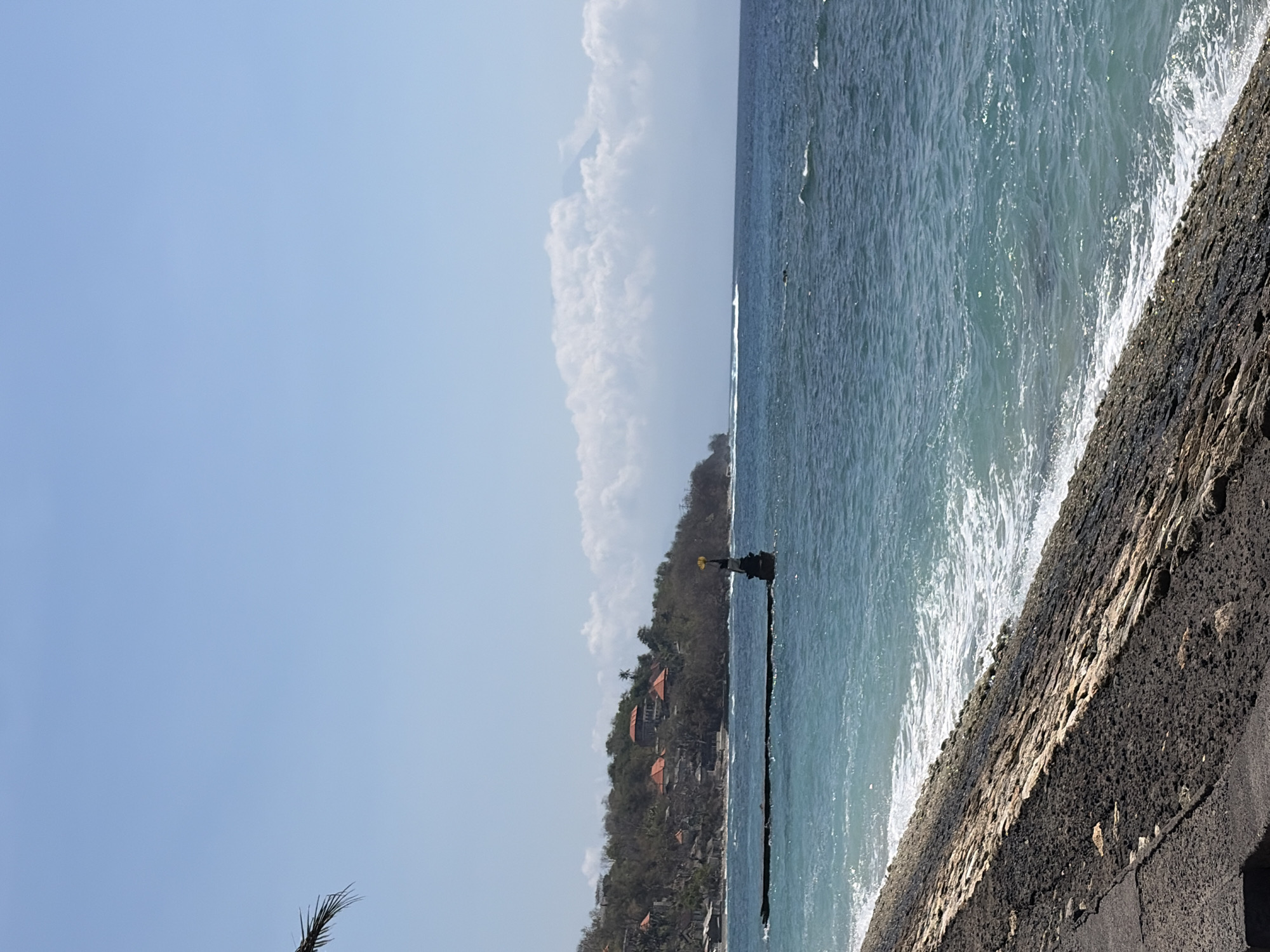 The image depicts a coastal scene with a stone embankment in the foreground. Beyond the embankment, the turquoise ocean stretches out, with gentle waves breaking on the shore. In the middle ground, a dark, elongated structure extends from the coastline into the water, with a small statue or monument on its end. Further inland, a lush, green hillside is dotted with buildings that have orange-tiled roofs. The sky above is a clear blue with a few wispy white clouds in the distance. The overall impression is one of a serene tropical location. The scene is likely located in Nusa Penida, Indonesia, a popular tourist destination known for its picturesque coastlines and vibrant marine life.