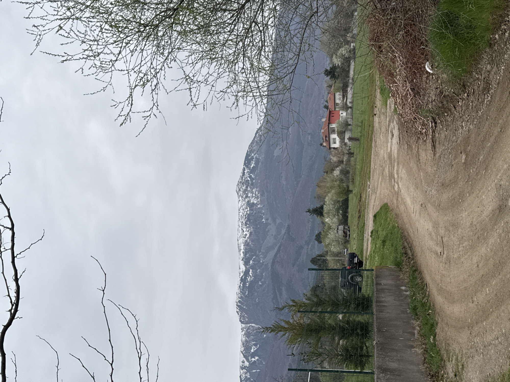The image shows a rural scene with a dirt road curving to the right. A mountain range, partially covered with snow, dominates the left side of the frame. A cluster of buildings, including a red-roofed house, is visible in the mid-ground to the right of the mountain. Branches of bare trees frame the top and bottom left corners of the image. A fence and evergreen trees are present in the foreground. A dark-colored vehicle is parked near the fence. The sky is overcast. The location is suspected to be in the vicinity of the city of Sofia, Bulgaria, based on the landscape and architectural style.