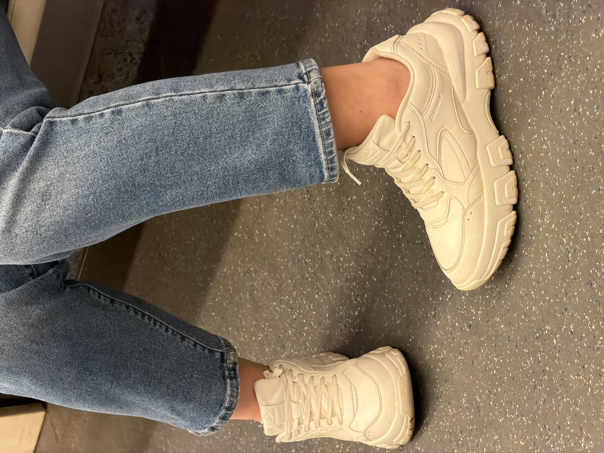 The image shows a pair of legs clad in light-blue jeans, terminating in white sneakers. The jeans are cuffed at the ankles. The legs are positioned on a gray, speckled surface. Based on the presence of the DEMIX logo on the sneaker, and the speckled floor often found in public transit, the location is most likely the Moscow Metro.