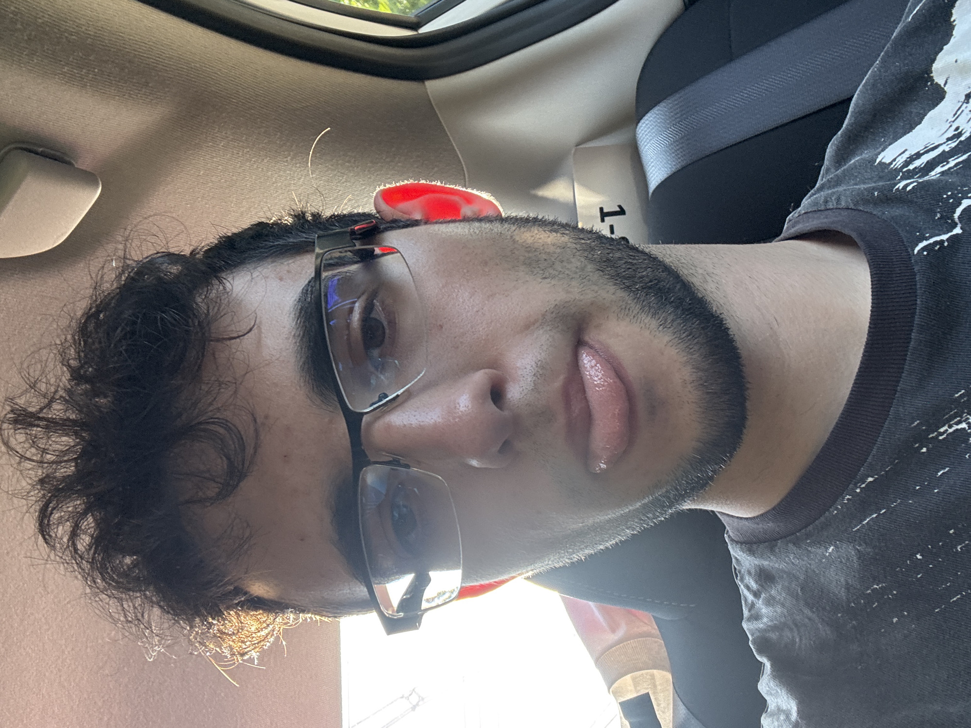 A selfie photograph depicts a person with dark curly hair, a beard, and glasses, positioned inside a vehicle. The person is wearing a dark-colored t-shirt. A seatbelt is visible in the background, along with interior car components. The location is specified as Franklin Township, United States of America.