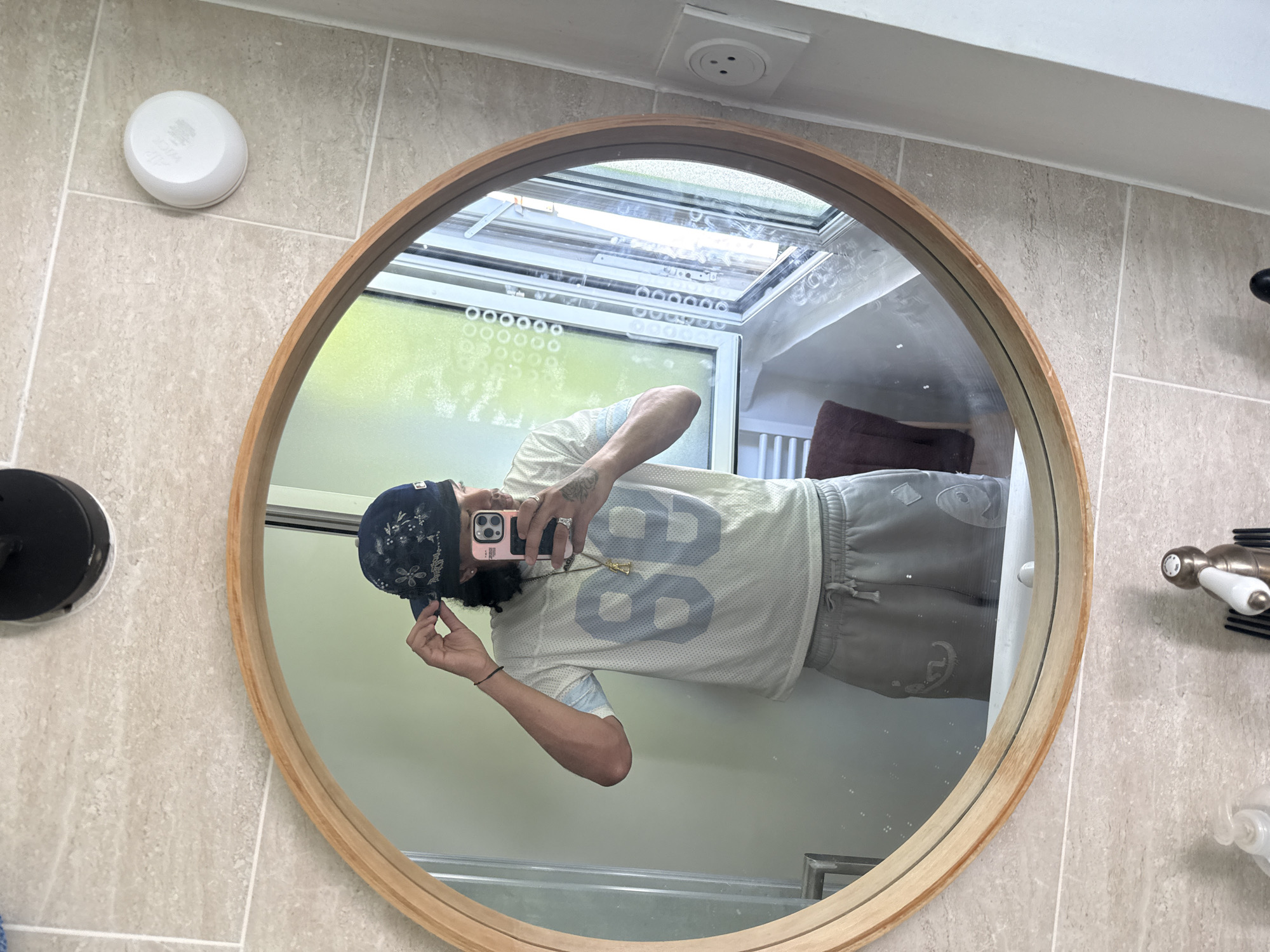A person is standing in front of a round mirror in a bathroom, taking a selfie with a cell phone. The person is wearing a dark patterned bandana, a light blue and white jersey with the number "88" on the front, and gray sweatpants. The bathroom has light-colored tiled walls and floor. Through the mirror, a window with blinds is visible, and a white electrical outlet is on the wall to the right. The image does not provide any information to determine the location in Massy, France.