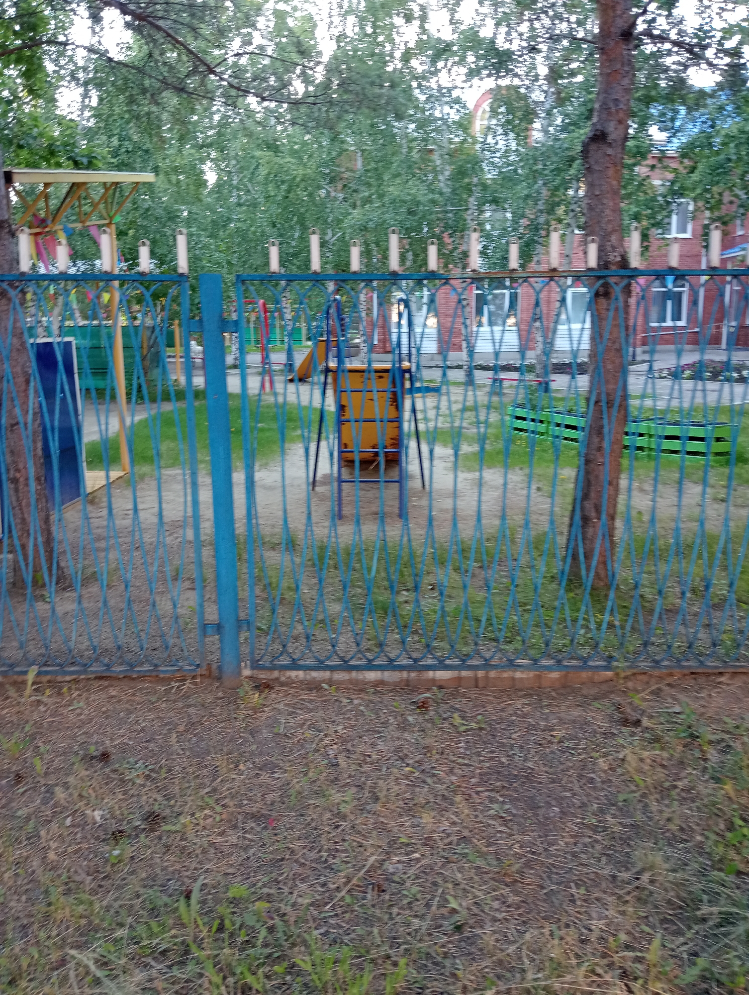 An outdoor medium shot shows a turquoise metal fence with a diamond pattern in the foreground, positioned in front of a playground. Behind the fence, elements of the playground are visible, including a yellow structure with a blue frame, green planter boxes, and a hint of a slide in the background. The environment appears to be a grassy area with scattered dry leaves and patches of green grass. Trees with mature trunks and green foliage are present on either side of the scene, and behind the playground, a brick building with windows can be seen. The lighting suggests it is daytime.