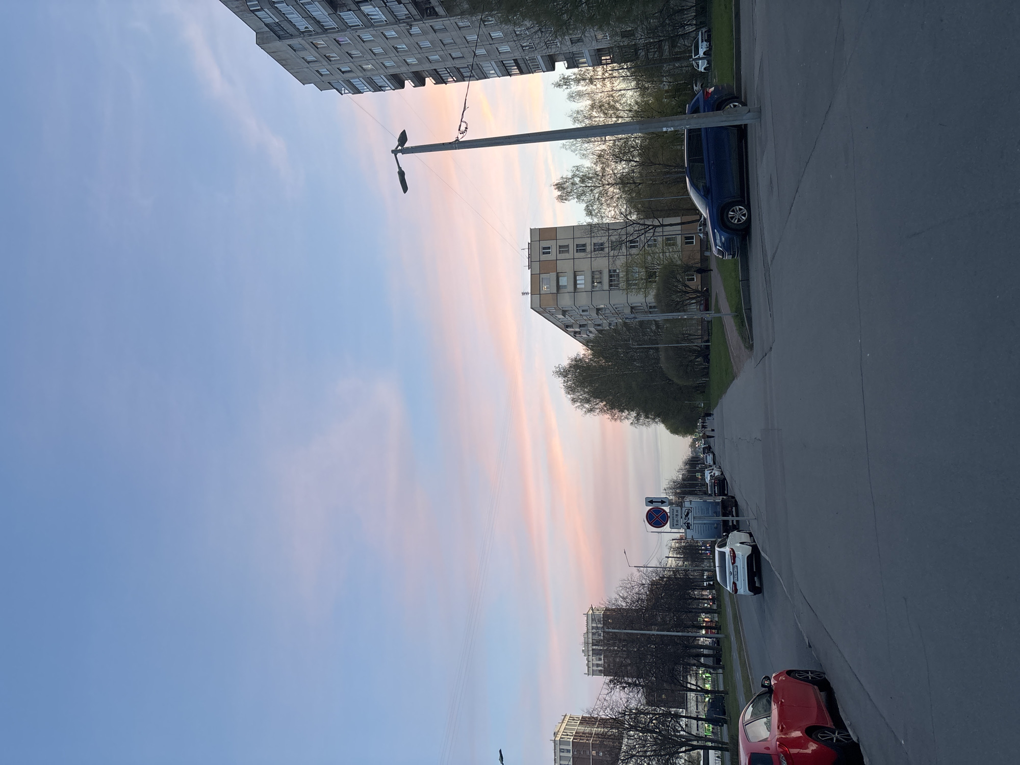 This media file captures an urban street scene at what appears to be either sunrise or sunset, indicated by the long, wispy streaks of pink, orange, and blue clouds across the sky. The image is taken from a slightly elevated perspective looking down a road that recedes into the distance.
On the left, a tall, multi-story residential building with numerous windows dominates the upper frame, appearing to be of a Soviet-era architectural style. Further down the street on the right, another similar apartment block is visible. The road is paved, and cars are parked along its right side. A prominent blue sedan or small SUV is parked in the mid-ground. Further down, a white car is parked near a complex traffic sign, and a red car is partially visible in the bottom right corner. Other vehicles can be seen in the distance, suggesting active traffic or parked cars along the entire stretch of the road.
A tall street light pole with multiple fixtures stands on the right side of the road, close to the blue car. The street is lined with trees, some with sparse foliage, indicating either early spring, late autumn, or a deciduous species. Patches of green grass are visible along the roadside and between the road and buildings.
A notable detail is a white rectangular sign with various traffic indicators. The most prominent is a circular "No Stopping" sign (red border, blue background, two crossed red lines). Below it, text reads "9:00 - 18:00," likely indicating time restrictions. Other Cyrillic text, partially obscured, appears to provide further details regarding parking regulations, potentially mentioning "кроме машин" (except for cars). The overall atmosphere is calm and quiet, characteristic of either early morning or late evening. The scene depicts a typical residential street in Vsevolozhsk, Russia, consistent with the architectural style and visible text.