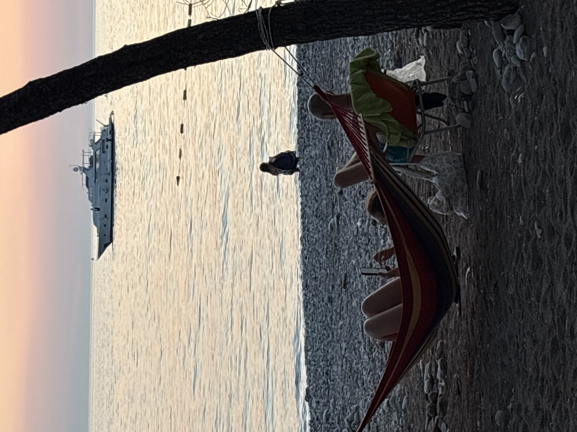 In Bar, Montenegro, at sunset, two people relax on a pebble beach. A woman reclines in a colorful hammock, looking at her phone. Beside her, a man sits in an orange chair with a green towel draped over it. In the background, a large white yacht is anchored on the calm sea, and another person is crouched near the water's edge. A tree trunk on the right frames the scene, with its bark textured and dark against the soft pastel hues of the sky and water. The overall atmosphere is tranquil and serene.