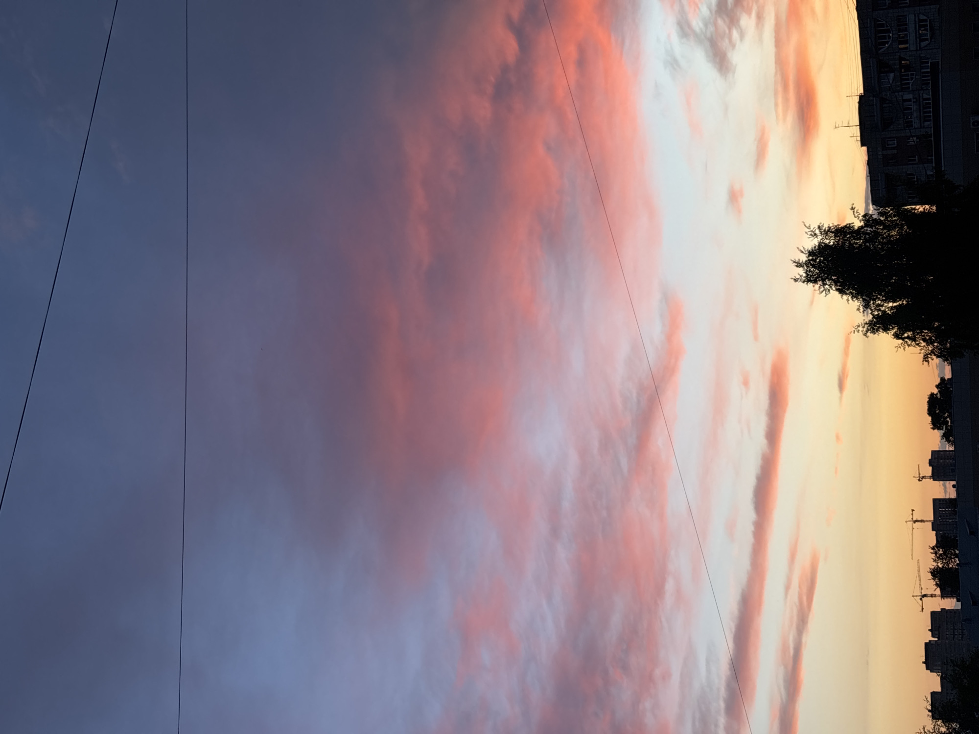 The image depicts a sunset in Perm, Russia. The sky transitions from a blue hue on the left to orange and pink on the right, with long, horizontal clouds tinted with pink. Power lines stretch across the sky. On the right, the silhouette of a building, trees, and construction cranes are visible.