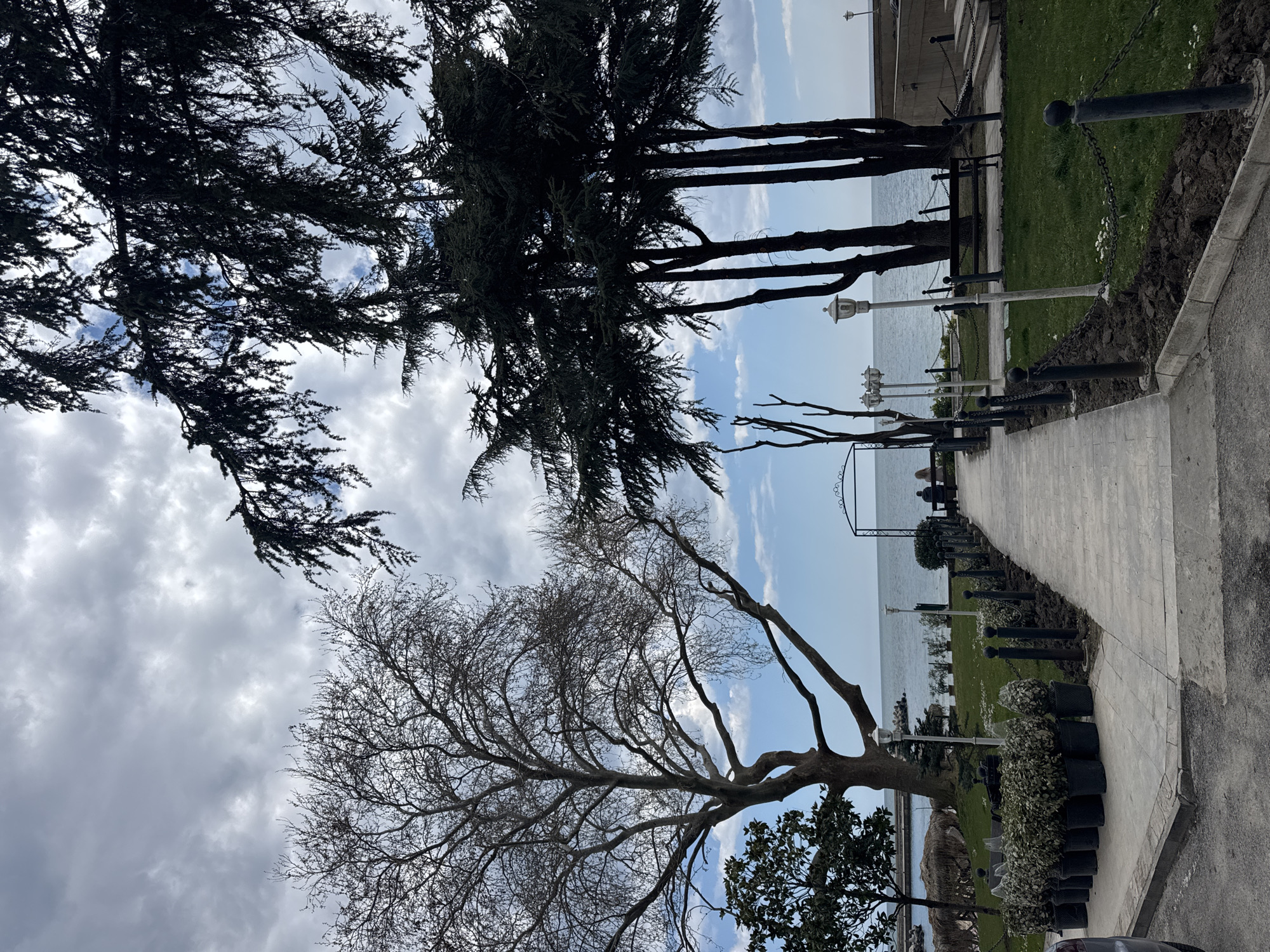 The image shows a park scene along the Black Sea coast in Varna, Bulgaria. A paved pathway runs along the coast, bordered by grass and trees. The sea is visible in the background, with a light blue sky and some clouds. A line of dark-colored metal posts with chains runs parallel to the path. The trees are a mix of evergreen and deciduous, the latter being bare. There are several potted plants along the path. There are two people sitting on a bench in the distance. The light poles are visible along the pathway.