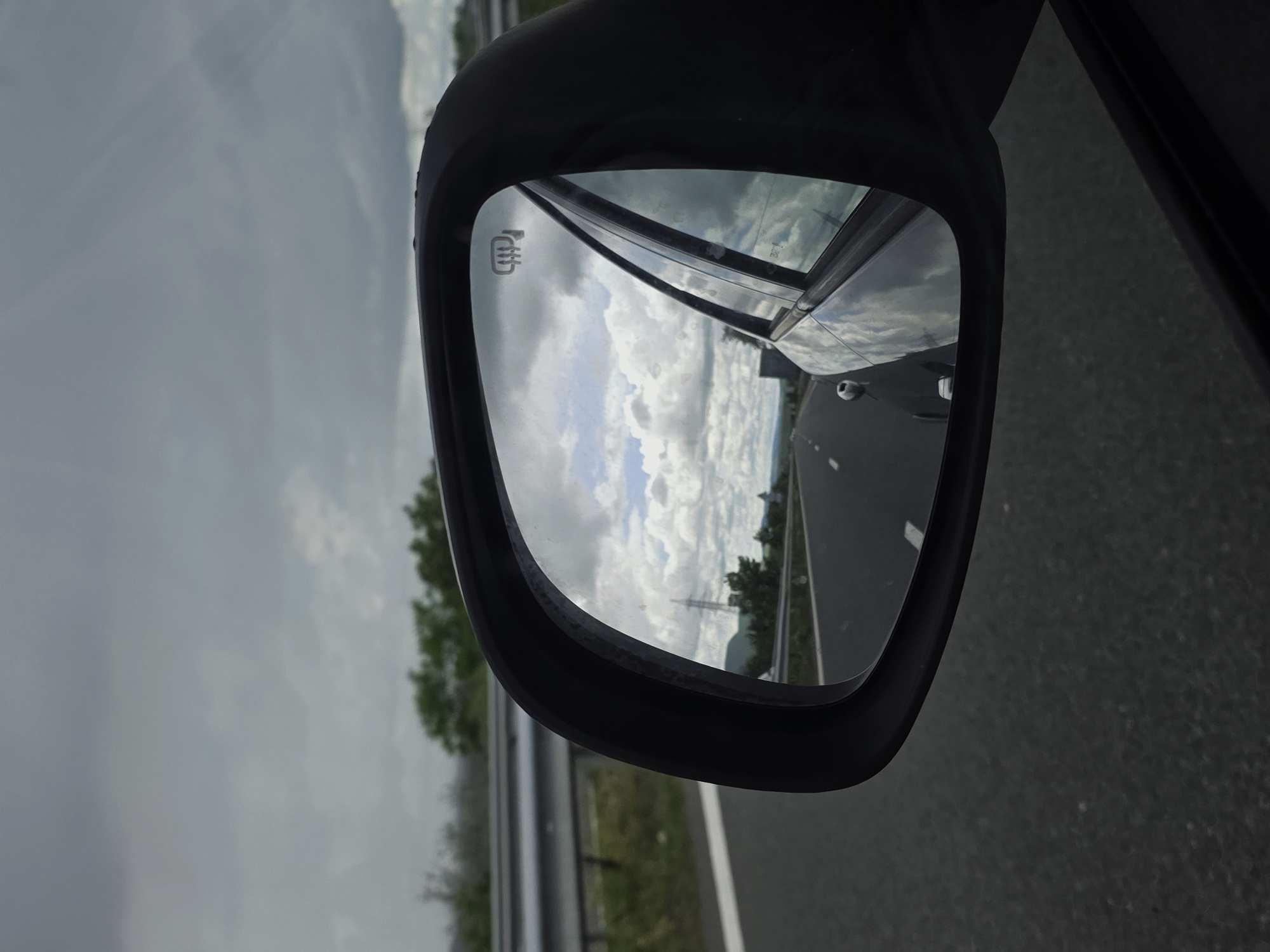 The image depicts a side-view mirror of a car while traveling on a road. The mirror's reflection shows another vehicle on the same road, along with a landscape featuring trees, mountains, and a cloudy sky. The car is traveling on a highway or major road. The exact location cannot be determined as it is a side view mirror of a moving vehicle. It is not possible to tell if the image was taken in Kaspichan, Bulgaria.