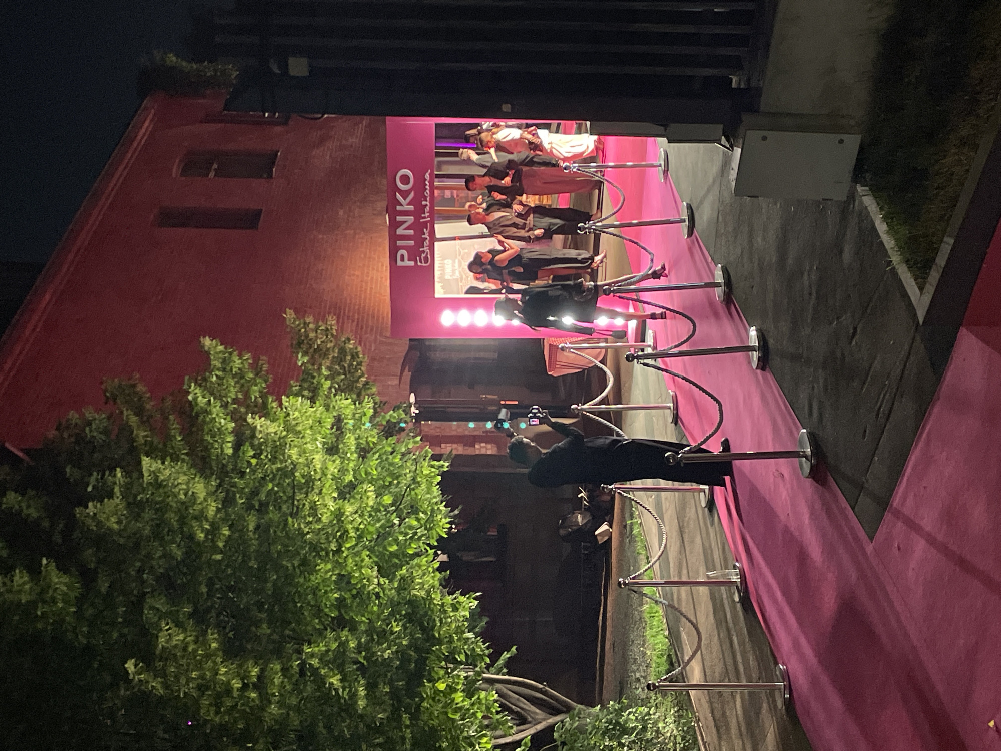 The image shows a nighttime event taking place at an establishment in Moscow, Russia. A pink carpet is arranged in front of a building, lined with chrome stanchions and ropes to guide people. A sign displaying "PINKO Estate Italiana" is present, indicating the event is associated with the fashion brand PINKO. Several individuals are gathered on a raised platform, illuminated by bright lights, with additional people visible in the background. A photographer stands along the carpet, using a camera to capture the event. The surrounding environment includes a building with dark red brick and a large, leafy green tree. A gray wall is located to the right of the pink carpet.