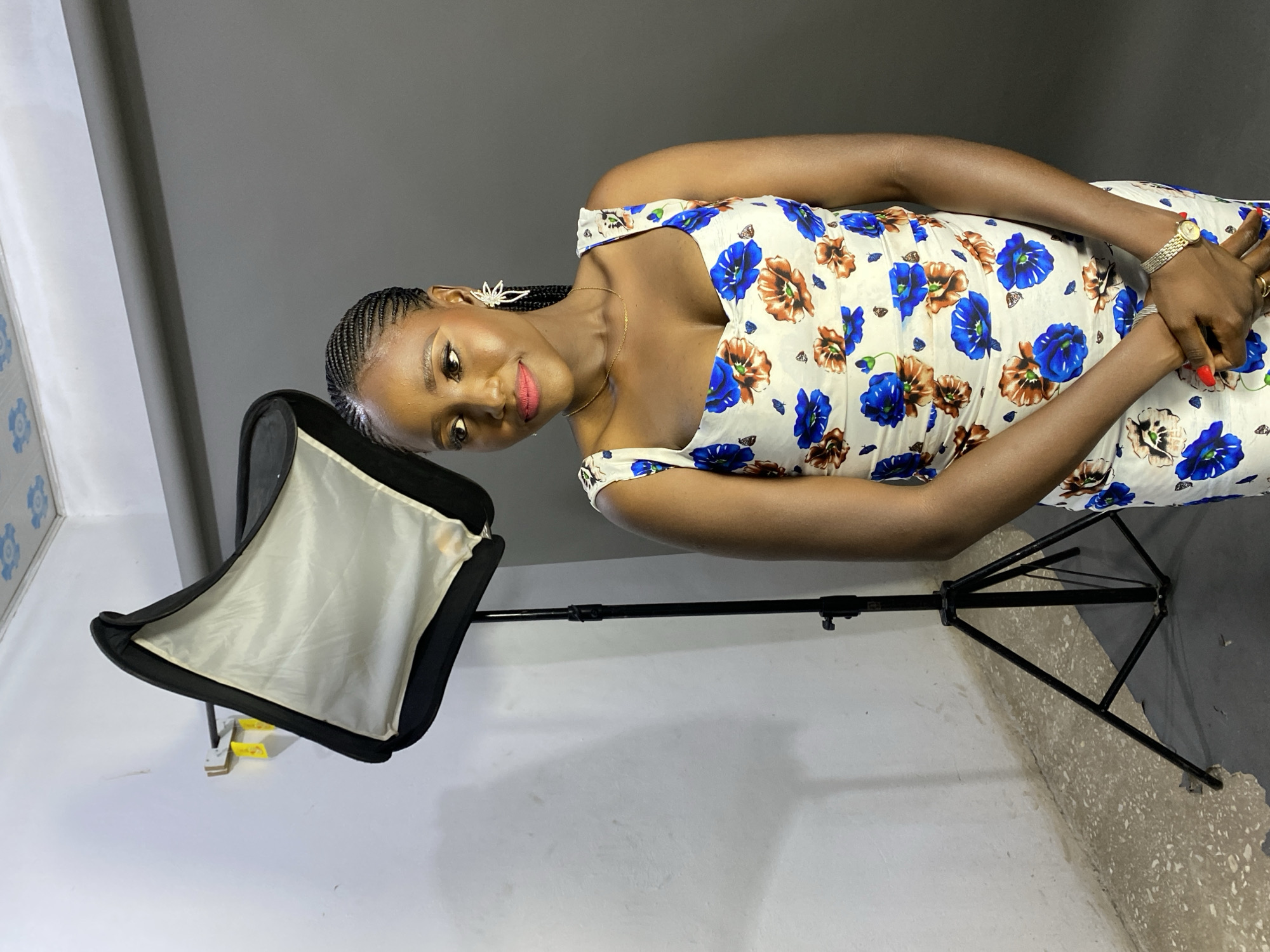 A young woman with dark skin is depicted in a portrait-style photograph, posing indoors in what appears to be a photography setup. She stands slightly angled, looking directly at the camera with a calm and subtle smile. Her hair is styled in neat braids, and she wears decorative, starburst-like earrings, a delicate gold necklace, and a gold-toned watch on her left wrist. Her fingernails are painted red, and she wears rings on her right hand, which is clasped over her left hand in front of her.
The woman is wearing a sleeveless or spaghetti-strap dress with a white base, intricately patterned with vibrant blue and earthy brown flowers, along with small dark butterfly motifs. The setting features a plain gray wall in the background, which meets a lighter, possibly white, wall on the left side of the frame. The floor transitions from white to a grayer, speckled surface. A black softbox light modifier, mounted on a tripod stand, is visible on the left, indicating artificial lighting for the photoshoot. The overall scene suggests a professional or semi-professional photography session taking place in Jos, Nigeria. There is no visible text in the image.