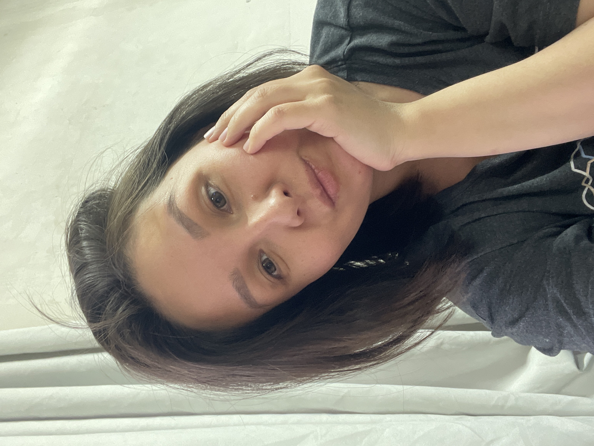 A woman is visible in the image. She has brown hair and is wearing a gray t-shirt. Her right hand is touching her face. The background appears to be a white wall or curtain. It is difficult to determine the exact location without additional information. The location is presumed to be in the city of Verkh-Katunskoye, Russia, based on the user's request, but there is no evidence of that in the image.