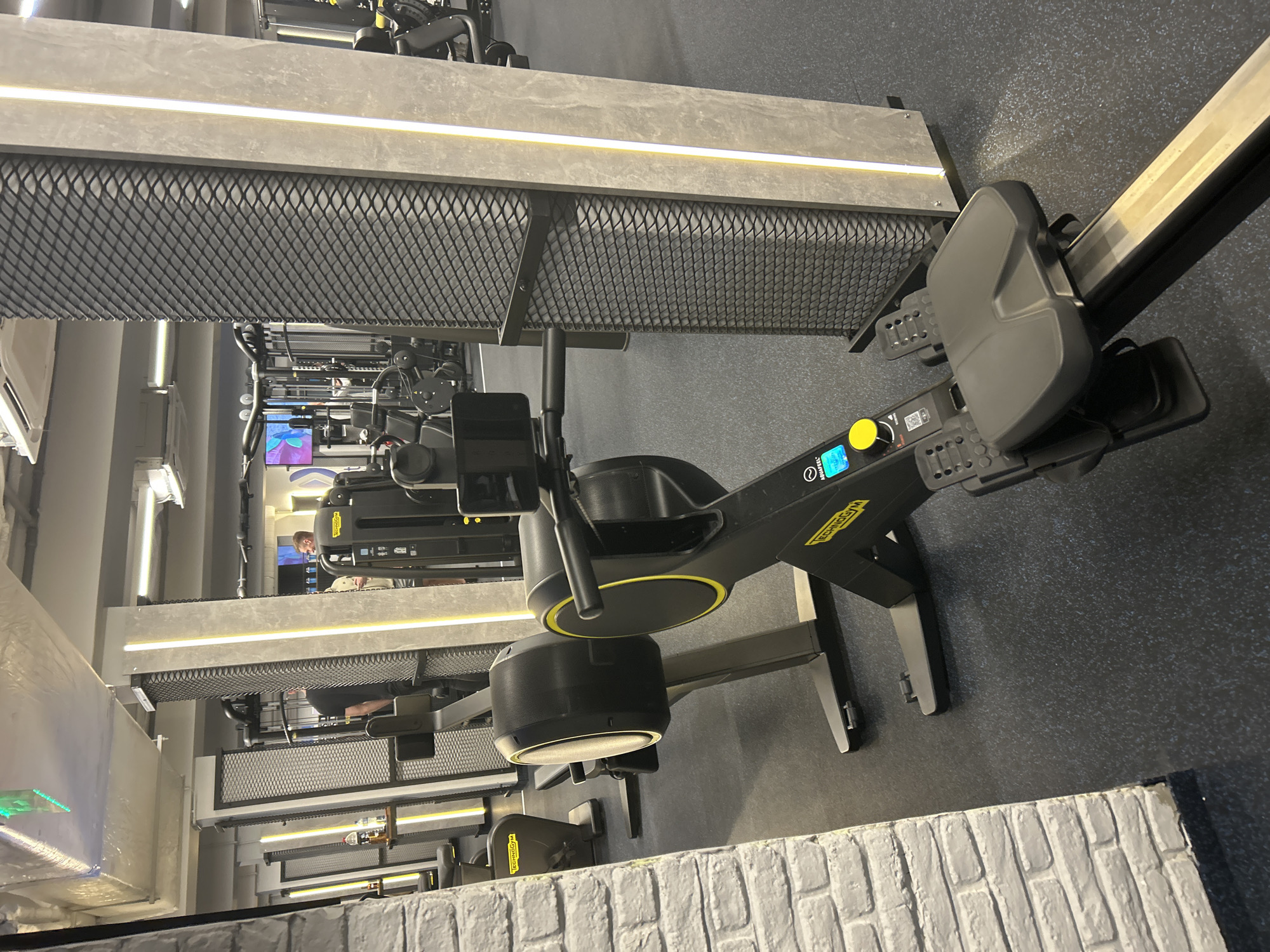 The image depicts a gym interior featuring multiple Technogym rowing machines. The machines are positioned in a row, with black frames and yellow accents. Above the machines are architectural elements consisting of concrete slabs with integrated LED lighting and a mesh grate. The flooring appears to be a dark blue or gray rubberized surface. In the background, other gym equipment and individuals can be seen, but they are out of focus. The foreground includes a faux white brick wall detail. The overall environment suggests a modern and well-equipped fitness facility. The location cannot be determined solely from the image.