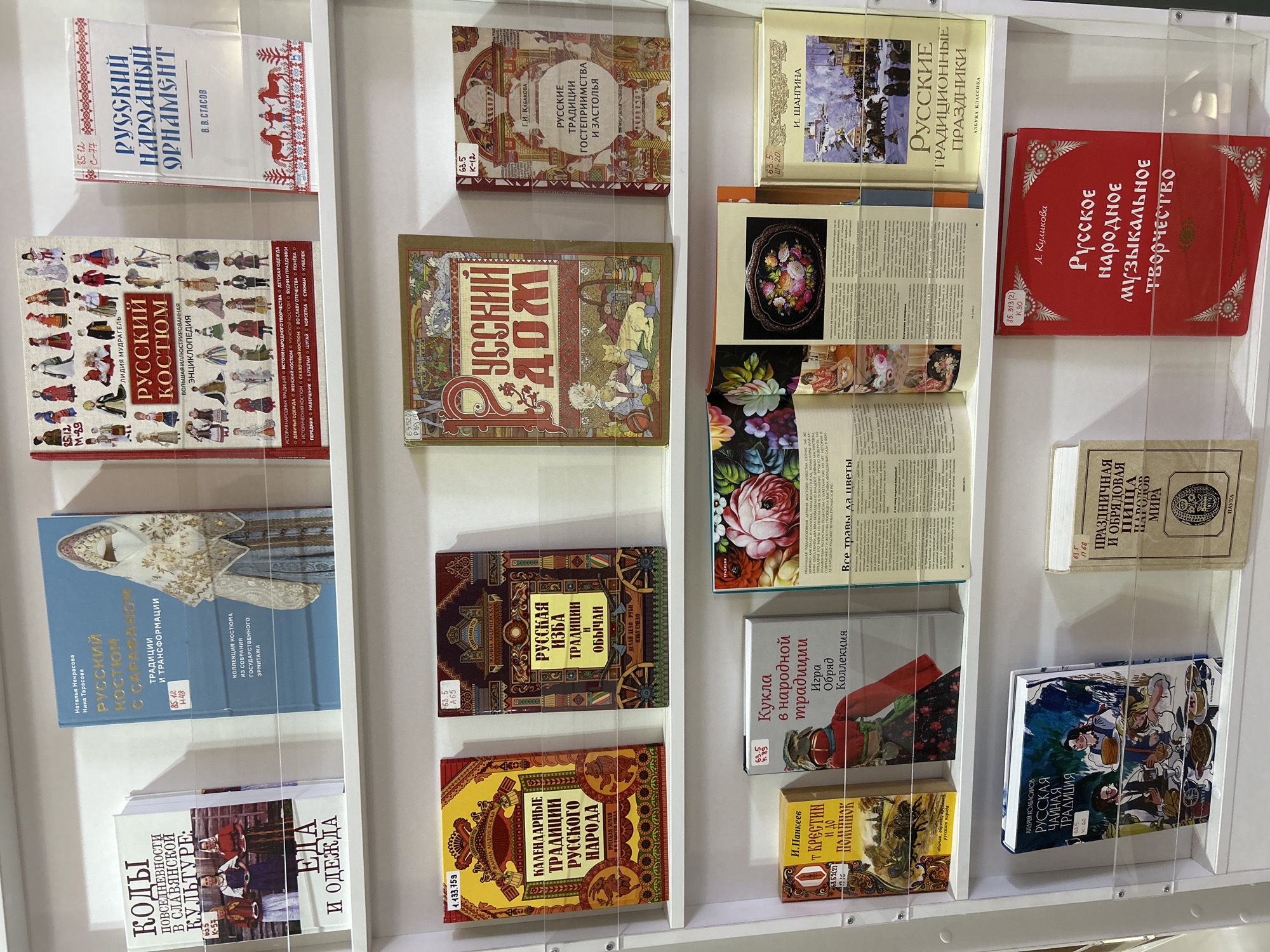 The image shows a bookshelf with several books on Russian traditions and culture. The books cover topics such as folk costumes, ornaments, calendar traditions, cuisine, and musical creativity. Some of the books are open, revealing colorful illustrations and text. The titles are in Russian, and the bookshelf is likely located in a bookstore or library.