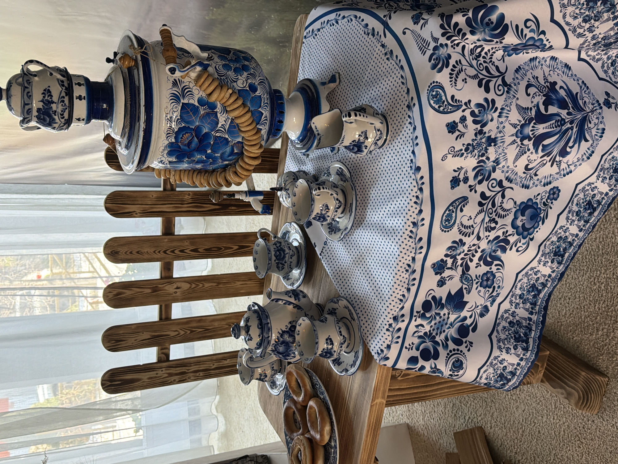 The image displays an indoor setting arranged for a traditional Russian tea ceremony, situated in Izhevsk, Russia. A rustic wooden table supports the arrangement. A white tablecloth, intricately patterned with blue Gzhel-style floral motifs and small blue dots, drapes over the table.
The central object is a large, ornate samovar, decorated with blue floral patterns characteristic of Gzhel porcelain. A smaller, matching teapot with a lid rests on top of the samovar. A garland of light brown, ring-shaped *sushki* or *baranki* hangs around the samovar's upper body.
To the left of the samovar, a matching blue and white Gzhel-style porcelain tea set is arranged on the tablecloth. This set includes a lidded teapot, four teacups, four saucers, a small creamer, and a lidded sugar bowl or similar container. On the far left of the wooden table, a blue and white patterned plate holds approximately six additional *sushki* or *baranki*.
In the background, a large window is visible, covered by translucent white curtains. Through the curtains, bare tree branches and a light-colored outdoor structure are discernible, indicating an exterior view under daylight conditions. Behind the table, a wooden structure resembling a short fence or chair back, composed of vertical wooden slats, is partially visible. The floor beneath the table is covered with a light-colored carpet or rug. No individuals are present in the scene.