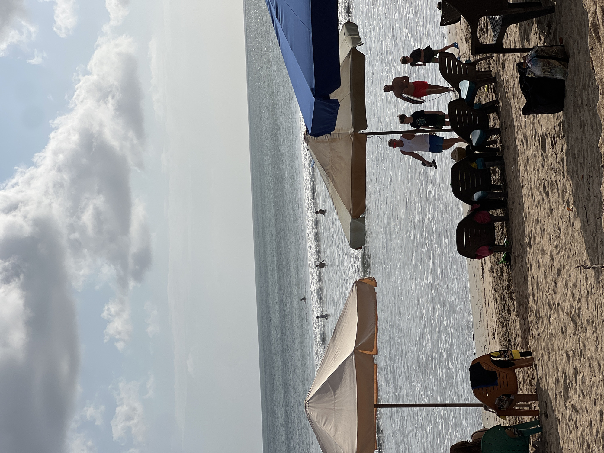 The image depicts a beach scene in Kuta, Indonesia. In the foreground, beach chairs are set up on the sand, some occupied by people and others empty. Several beach umbrellas are erected, providing shade. The middle ground shows the ocean with gentle waves and several people in the water, some appearing to be swimming or surfing. In the background, the horizon is visible where the ocean meets the sky, which is partly cloudy. A group of people are walking near the water's edge, one carrying a surfboard. The sand appears light brown and textured with footprints and debris. The overall atmosphere is relaxed and typical of a beach resort.