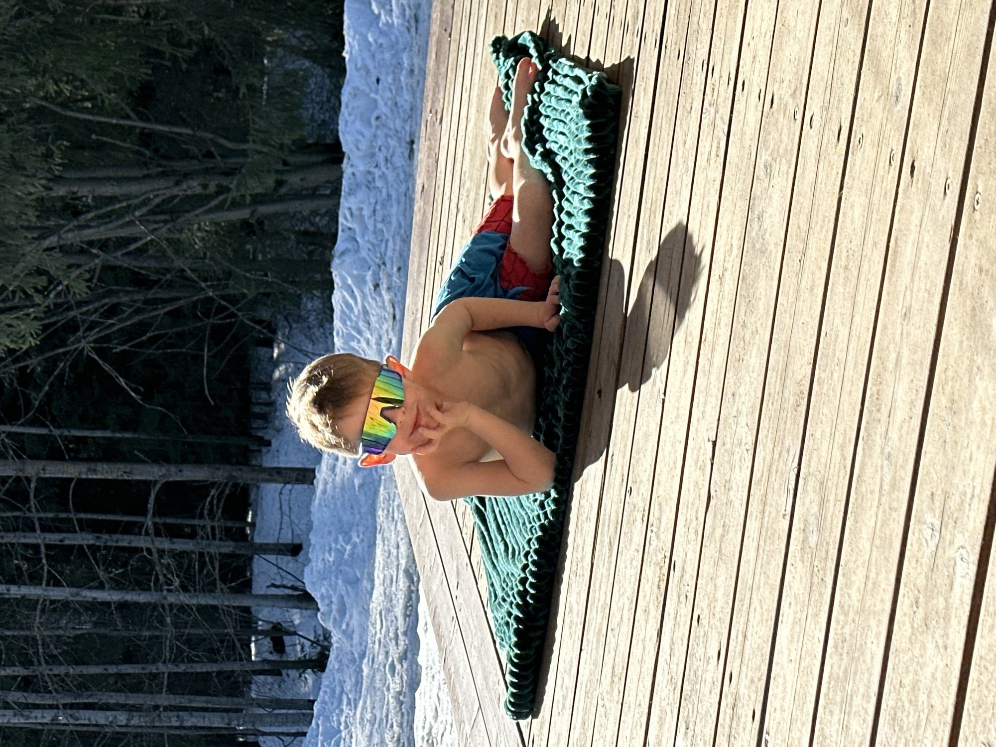A young, fair-skinned male child is depicted lying prone on a dark green textured blanket positioned on a light brown wooden deck in Прикамский, Russia. The child, appearing shirtless and wearing red and blue patterned shorts, has light-colored hair and is wearing reflective, multi-colored athletic sunglasses. His right hand supports his head, with his elbow resting on the blanket, while his left arm is bent, hand near his chest. He faces the camera with a subtle expression. Strong sunlight illuminates the scene, casting distinct shadows on the wooden deck. Immediately behind the deck, the ground is covered in patches of snow, which transitions into a dense background of trees, including both bare deciduous and evergreen varieties, consistent with a wooded area.