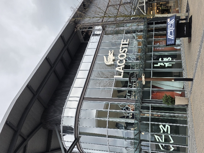 The image depicts the exterior of a retail store, identified as "LACOSTE," situated within the Zweibrücken Fashion Outlet in Zweibrücken, Germany. The building features a modern design with extensive glass curtain walls forming the lower two levels, including a prominent curved corner section on the left. The upper section and roof are constructed from dark gray, possibly metal, cladding with visible structural beams supporting an overhang.
The "LACOSTE" brand name, in white capital letters, along with its crocodile logo, is prominently displayed on the glass facade. Below this, and on the curved glass section to the left, additional white neon-style lettering or patterns are visible through the glass.
In the paved foreground, composed of light-colored interlocking pavers with dark linear accents, several objects are present. A tall, light-colored planter containing a small green bush stands near the entrance. To its right, a dark brown, multi-directional signpost with a black base and an integrated light fixture extends several arms. Further to the right, a black digital kiosk displays a blue and white map, likely a directory for the shopping complex. A leafless deciduous tree with thin branches is visible on the far right. The sky overhead is overcast and light gray. No individuals are present in the scene, and no specific actions or interactions are depicted.