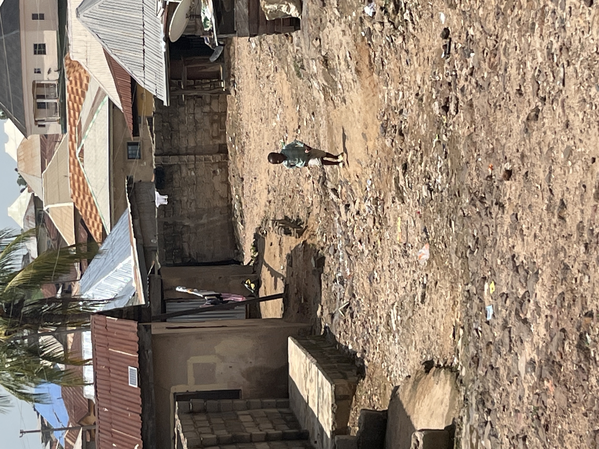 In an urban environment, a young child runs through a dusty, debris-strewn path. The child is wearing a light blue long-sleeved shirt and grey shorts, with yellow shoes. The background is filled with a dense collection of residential buildings, characterized by corrugated metal roofs of various colors, including red, silver, and orange. A palm tree with lush green fronds is visible on the left side of the image, partially obscuring the sky. The buildings are constructed from materials like stone and concrete blocks. In the foreground, the ground is covered in dirt, small rocks, and scattered litter, suggesting a well-worn path. To the left, a concrete structure extends from a building. The scene depicts a sunny day with visible shadows.