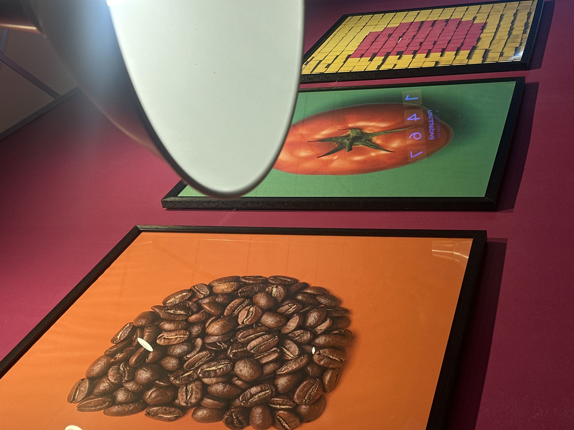 Three framed artworks are displayed vertically on a deep reddish-purple wall in an indoor setting in Votkinsk, Russia. A large, reflective, white-grey light fixture partially obscures the top left of the image, including part of the uppermost artwork.
The topmost visible artwork features a pixelated design composed of yellow and pink/red squares, possibly forming an abstract pattern or stylized shape. Below this, the middle picture prominently displays a ripe red tomato against a vibrant green background. Overlaid on the tomato are two distinct pieces of text: vertically oriented on the right side, the word "VeAL" is visible. Below it, inverted and in a stylized font, the word "rebellion" is discernible. The lowest artwork showcases a pile of roasted coffee beans arranged on a solid orange background.
The scene suggests an interior space, possibly a cafe, restaurant, or modern kitchen, given the food-themed art. Artificial lighting is implied by the presence of the light fixture. No people are visible, and no specific activity beyond the display of art is taking place. The overall impression is one of contemporary decor.