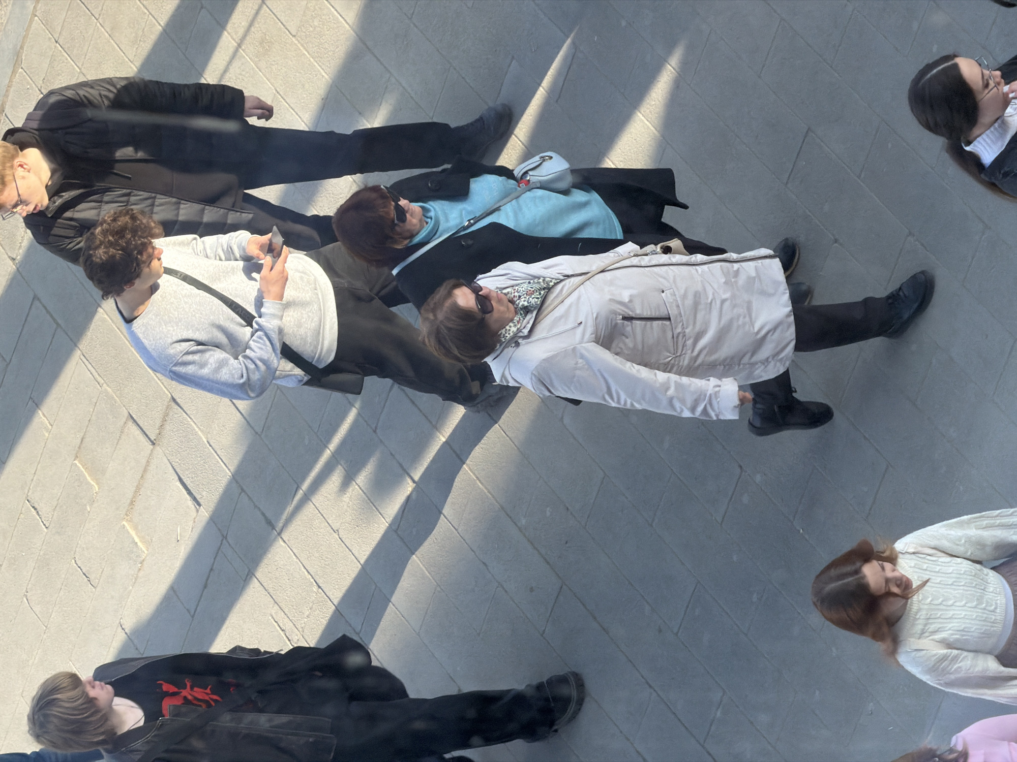 A top-down shot captures a group of people walking on a paved surface, possibly in Perm, Russia, indicated by the architectural style and pedestrian traffic observed.
The people, likely pedestrians, appear diverse in age and attire, with some wearing coats and jackets, sunglasses, and carrying bags. A person in a light gray sweater holds a phone.
The environment is characterized by a paved surface with shadows suggesting a sunny day. The paving pattern is geometric.