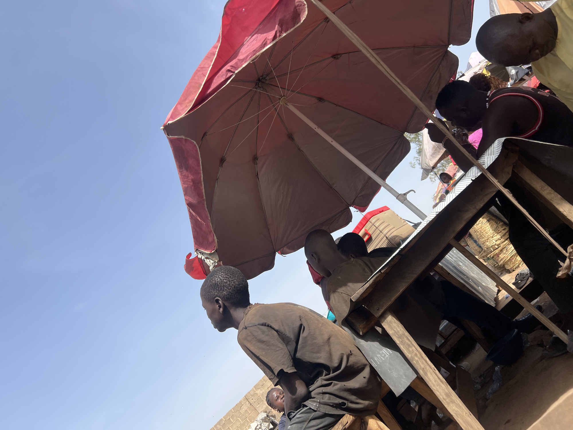 The image captures an outdoor scene from a low-angle perspective, looking upwards at a gathering under a large umbrella on a bright, sunny day. A significant portion of the frame, particularly the left side, is filled with a clear, light blue sky.
The central subject is a weathered red and brown umbrella, providing shade. Its underside, revealing the metal frame and spokes, is prominent in the upper middle of the image. Several individuals, primarily men and young people, are gathered beneath this umbrella. In the immediate foreground, a young man wearing a dark brown, possibly worn, t-shirt is seated, looking intently towards the left out of the frame.
Behind him and to the right, other people are visible, including a man in a dark tank top with red and white trim, and the head of another individual in a light-colored top at the far upper right. These individuals are situated around and within a rustic wooden structure, which appears to function as a stall or seating area. Various items like woven baskets and pieces of metal sheeting are visible within this structure.
The bright sunlight and the need for shade provided by the umbrella indicate it is daytime, likely midday or early afternoon, with clear weather. The setting appears to be an active outdoor space, possibly a market or a public gathering point. Faint white text, possibly "NEW-UP", is partially visible on a dark piece of material attached to the wooden structure in the lower right. The overall atmosphere suggests daily life and activity in a communal setting under the open sky.