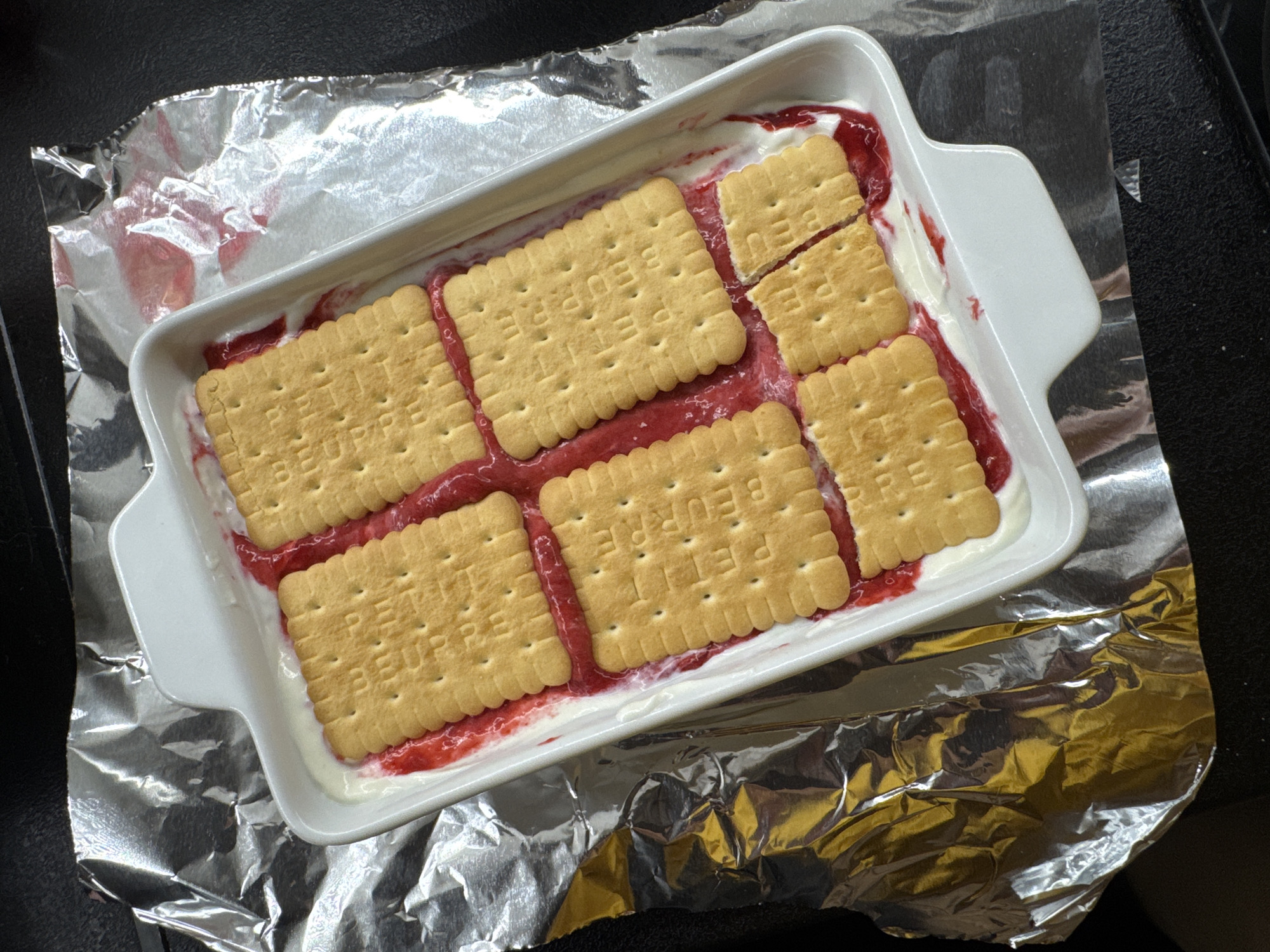 A top-down, eye-level shot shows a white rectangular baking dish filled with a dessert. The dish contains a white creamy layer topped with a red fruit compote and square biscuits. The biscuits are arranged in two rows of three on top of the compote, with some of them broken. The baking dish is placed on crumpled aluminum foil. The background is dark and out of focus. The location is Unknown, Unknown.