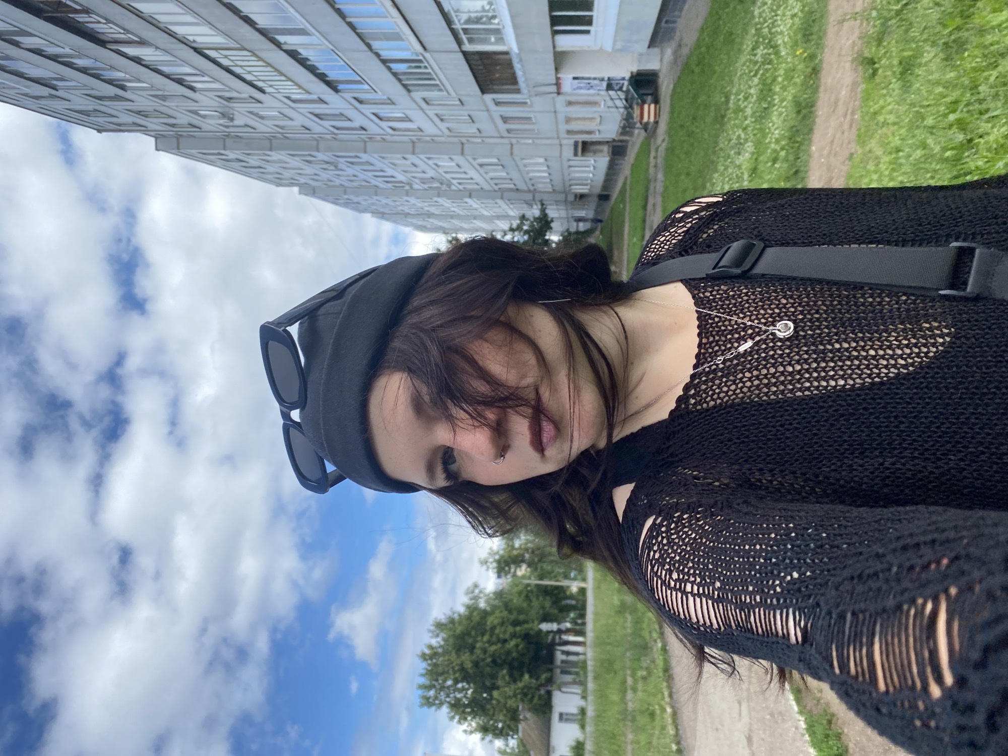 A young person with dark, wavy hair, wearing a black beanie with sunglasses perched on top, a black distressed knit top, and a silver necklace with a pendant, stands outdoors. They are looking to their right, with their hair partially obscuring their face. A black strap from a bag is visible across their chest.
The background features a tall, light-colored apartment building with multiple windows, typical of Soviet-era architecture, alongside a green grassy area with scattered white flowers. To the left, a less distinct building and trees are visible, suggesting an urban or suburban environment. The sky is partly cloudy with a mix of blue sky and white clouds. The scene appears to be located in the city of Chaikovsky, Russia, judging by the architectural style of the building and the overall setting. The lighting suggests it is daytime.